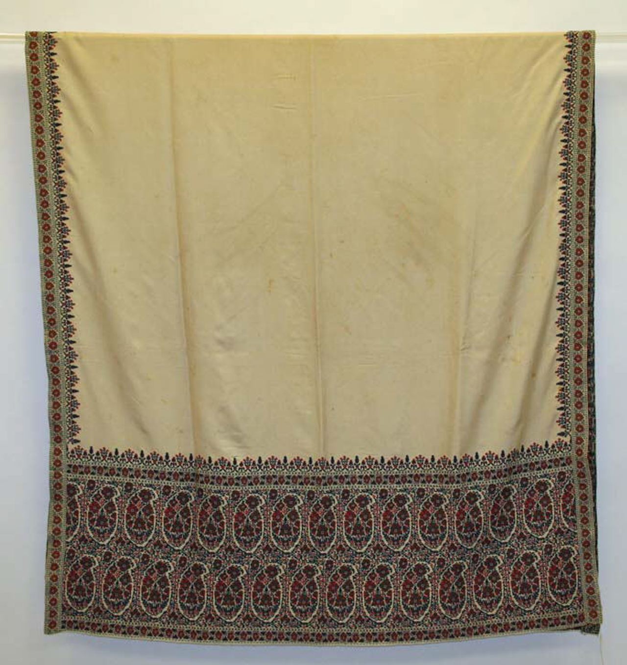 A cream-colored shawl hung over a rail, plain in the center, with narrow patterned side borders and a deep end border of rows of small red, black and white teardrop motifs.