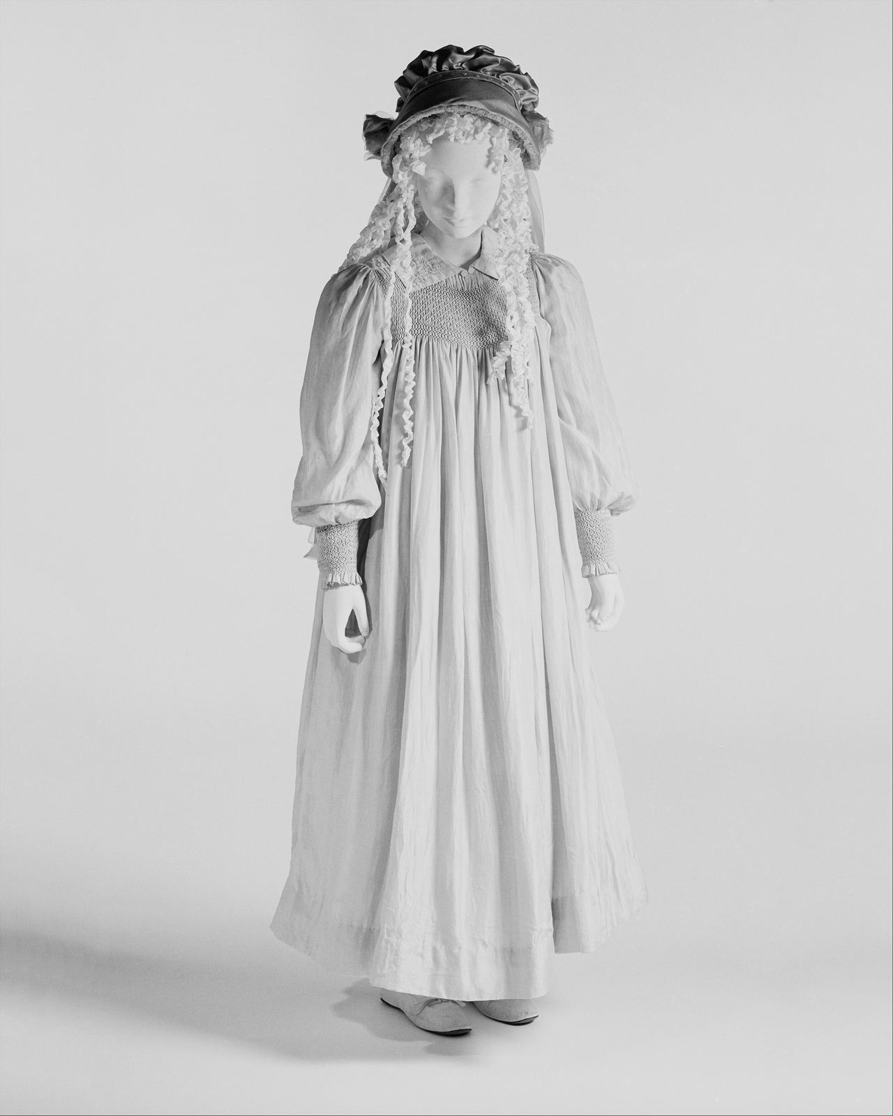A full-length pale dress of the 1890s on a child-sized mannequin, with a small smocked panel at the chest and smocked cuffs gathering the long sleeves.