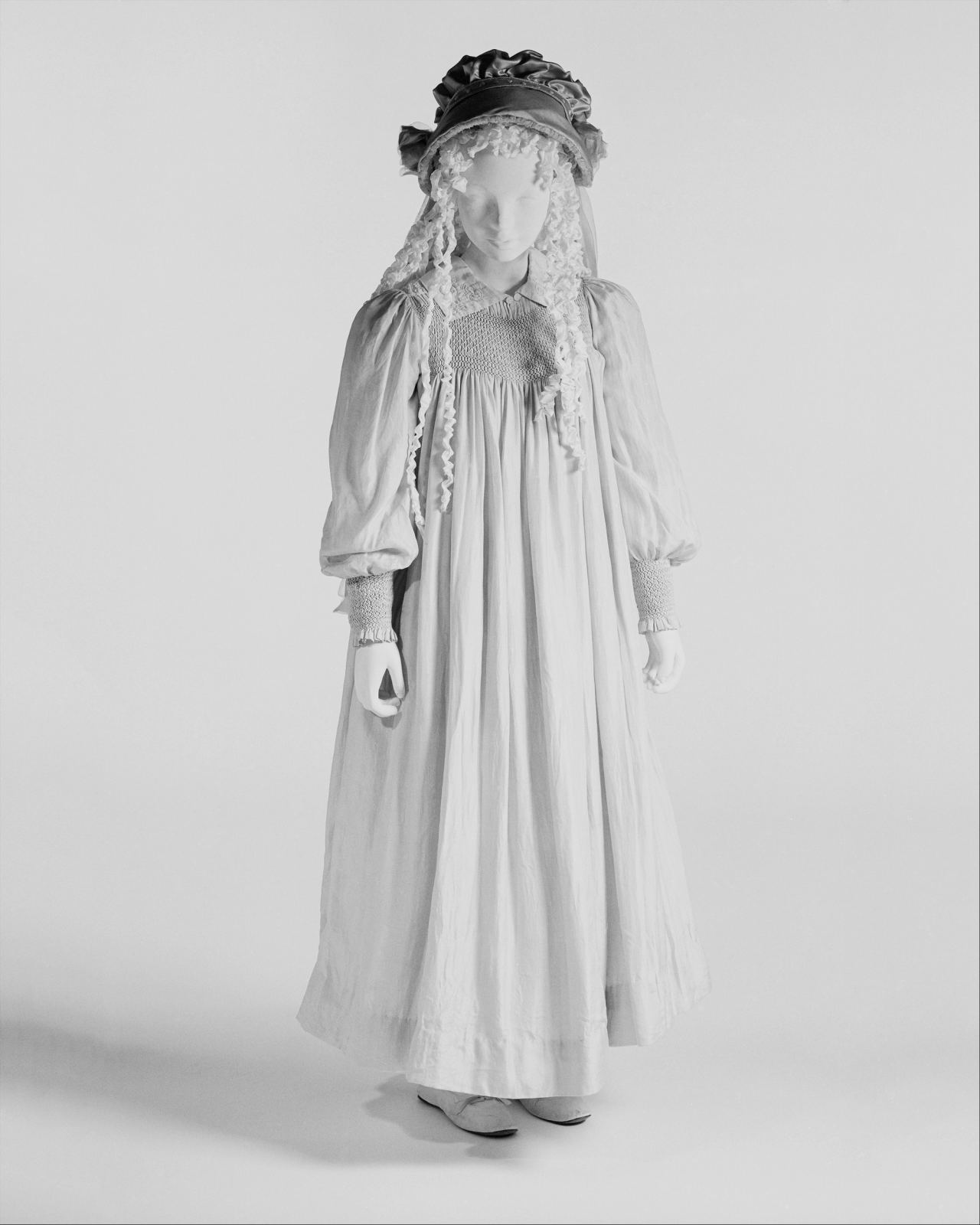 A full-length pale dress of the 1890s on a child-sized mannequin, with a small smocked panel at the chest and smocked cuffs gathering the long sleeves.