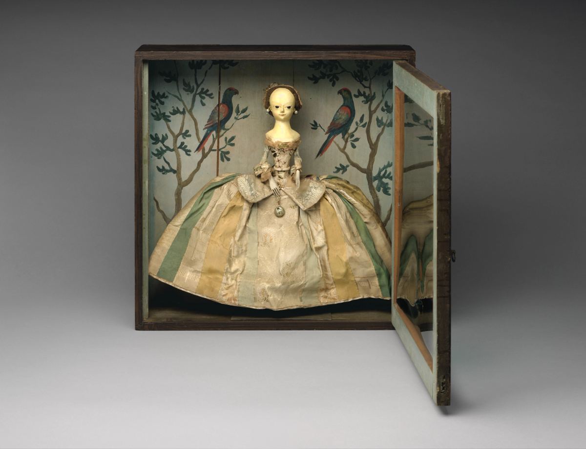 A wooden doll in a silk gown with a very wide skirt of green and cream stripes stands in an open wooden box. The box interior is painted with branching trees and two parrots.