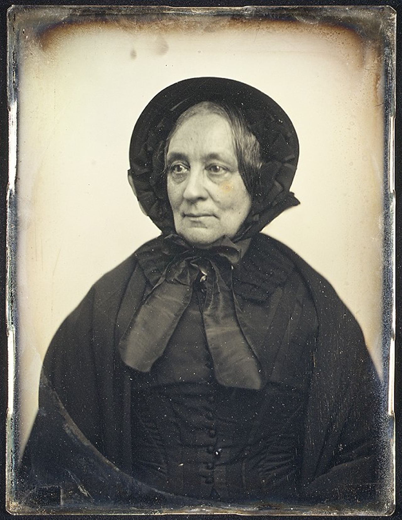 Daguerreotype portrait of an elderly woman looking to the left, in a black bonnet tied under the chin, a dark cape and a buttoned bodice with a large bow at the neck.