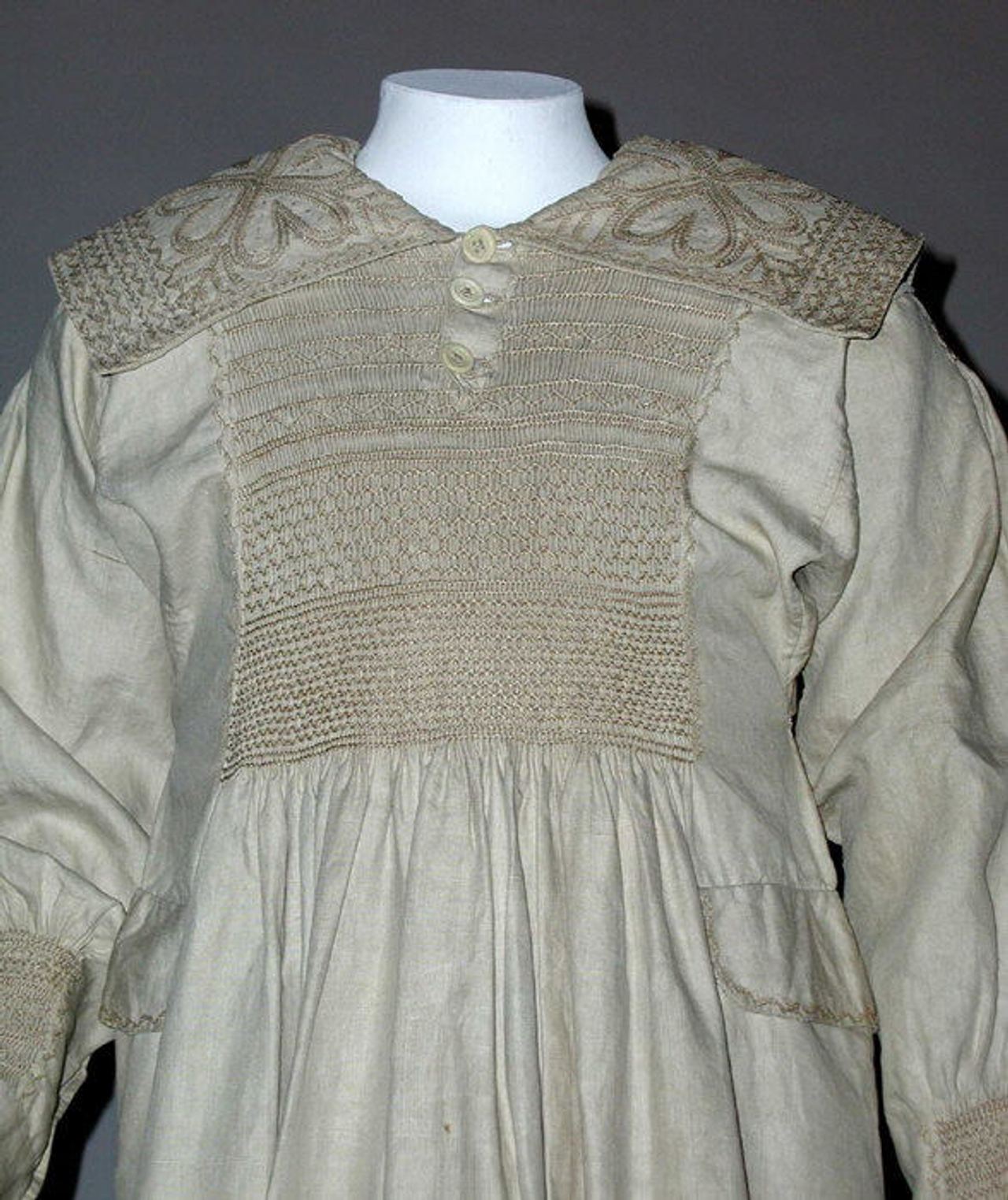 Close view of the smock's chest: rows of smocking, some forming a honeycomb-like lattice, between a wide collar worked with a flower motif and two buttons at the neck.