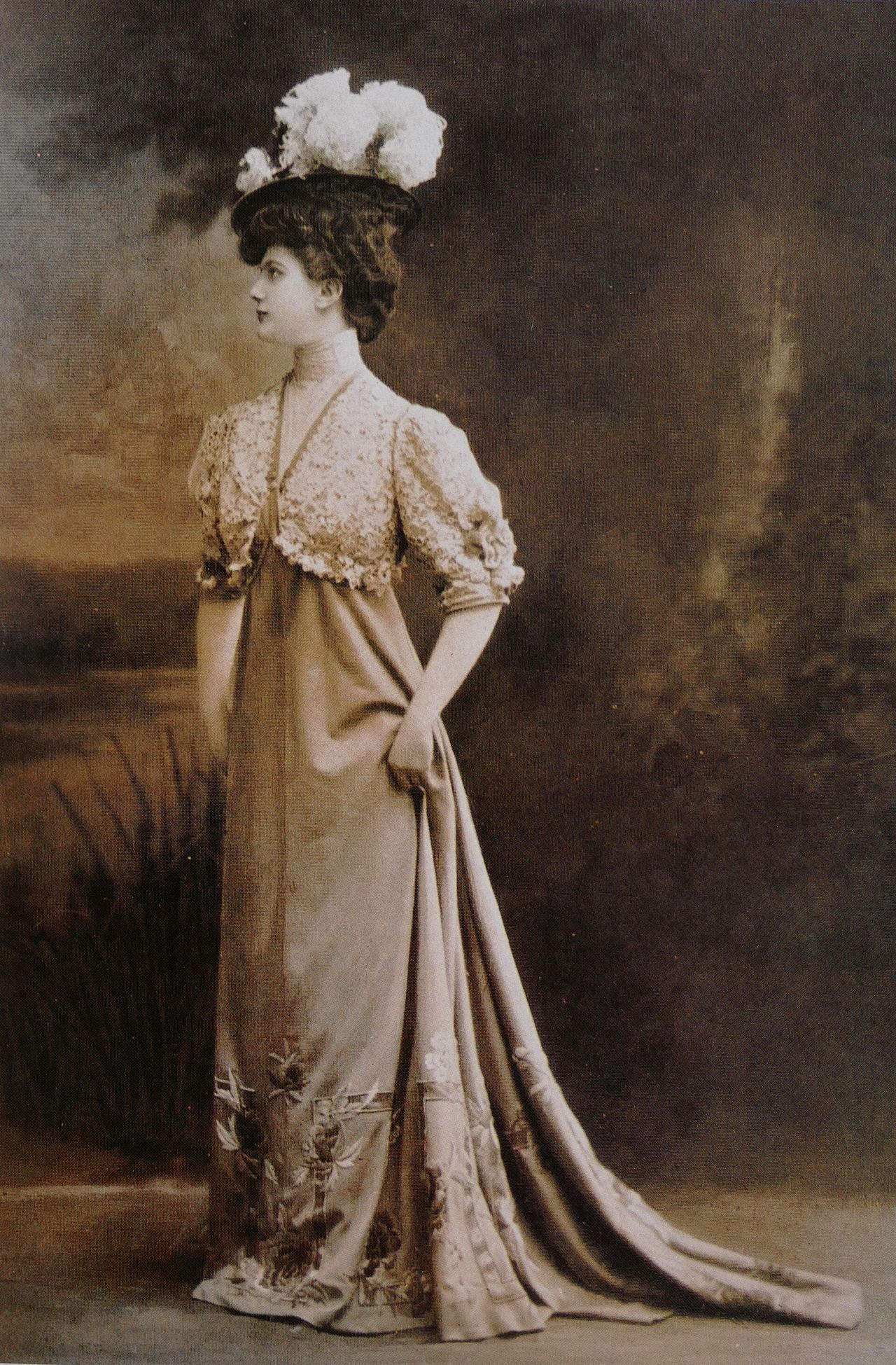 Sepia studio photograph of a woman in profile in a feathered hat and a long Paquin dress: a lace bodice with puffed elbow sleeves, a high waist, a plain skirt embroidered with flowers and lattice motifs at the hem, and a long train held in one hand.