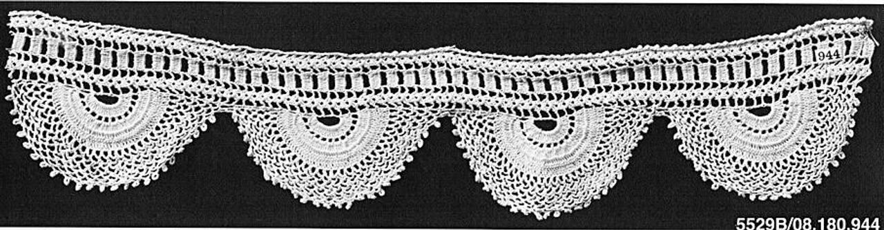 A black-and-white photograph of a white crochet border. A straight band along the top has rows of small rectangular openings; below it hang four half-round, fan-shaped scallops with a small knobbed edge.