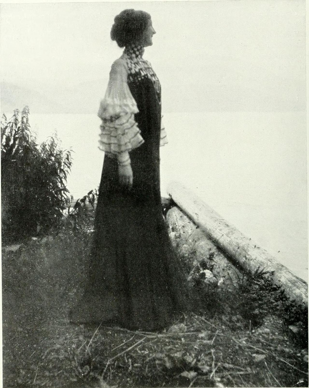 Black-and-white photograph of a woman seen in profile, standing at a lake shore beside a pale log and leafy shrub. She wears a long dark dress with a patterned high collar and yoke and white sheer sleeves finished in tiers of frills.