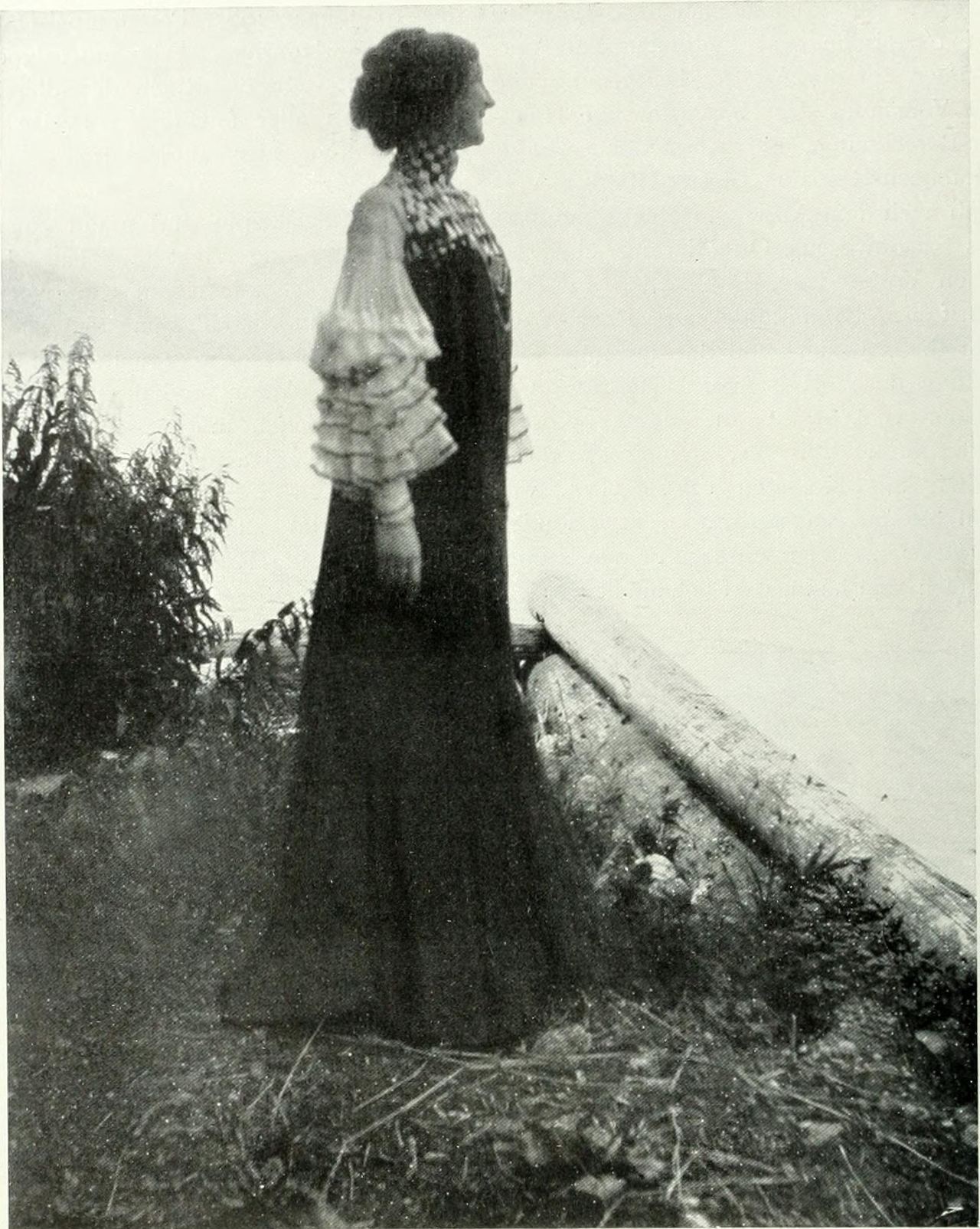 Black-and-white photograph of a woman seen in profile, standing at a lake shore beside a pale log and leafy shrub. She wears a long dark dress with a patterned high collar and yoke and white sheer sleeves finished in tiers of frills.