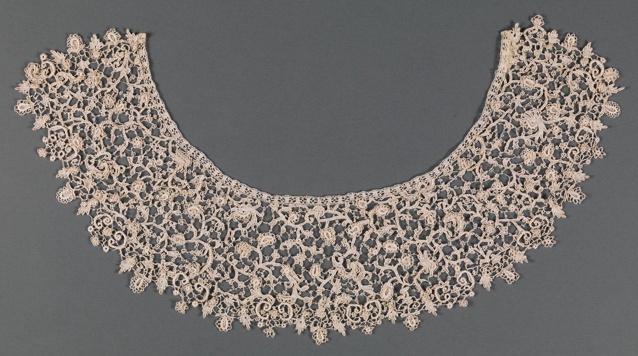 A wide, curved collar of cream needle lace on a gray ground: raised scrolling stems with leaves and flowers, tiny loops along the stems, and a narrow flat band of openwork along the inner neck edge.