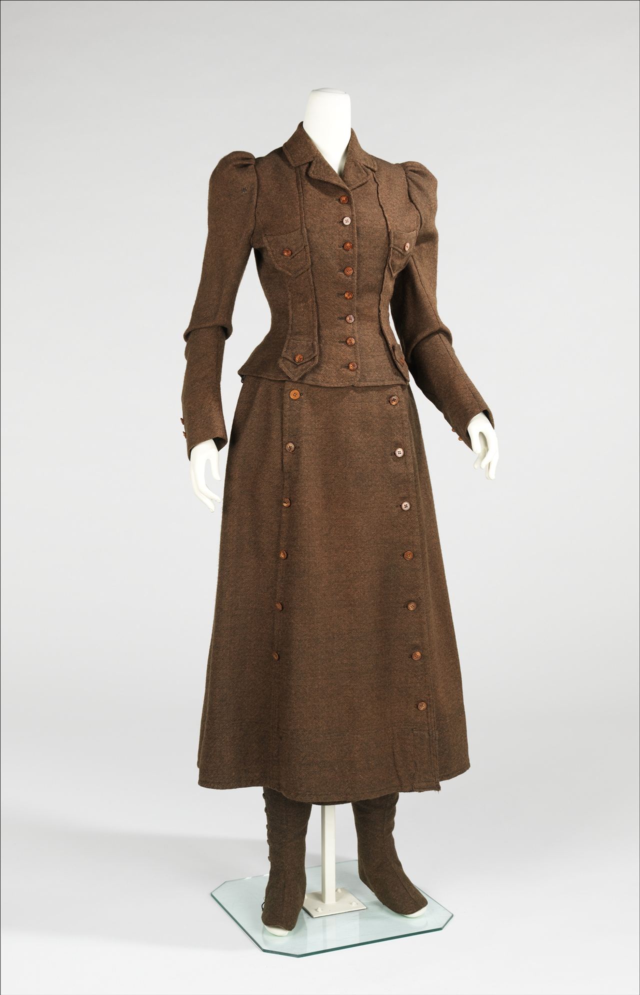 A brown wool jacket with puffed sleeves, flap pockets and a nipped waist, worn over a long plain skirt that fastens with two rows of buttons down the front; the tops of buttoned brown leggings show below the hem.