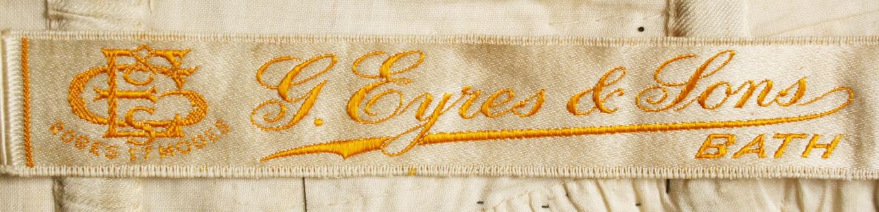A cream woven ribbon label with a gold monogram at left and the words G. Eyres and Sons, Bath in woven script, with the small words Robes et Modes beneath the monogram.