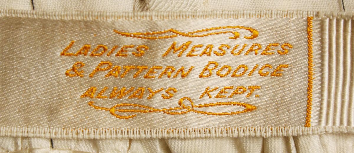 Close view of a cream woven ribbon label stitched inside a garment, with gold woven lettering that reads Ladies Measures and Pattern Bodice Always Kept, framed by small woven flourishes.