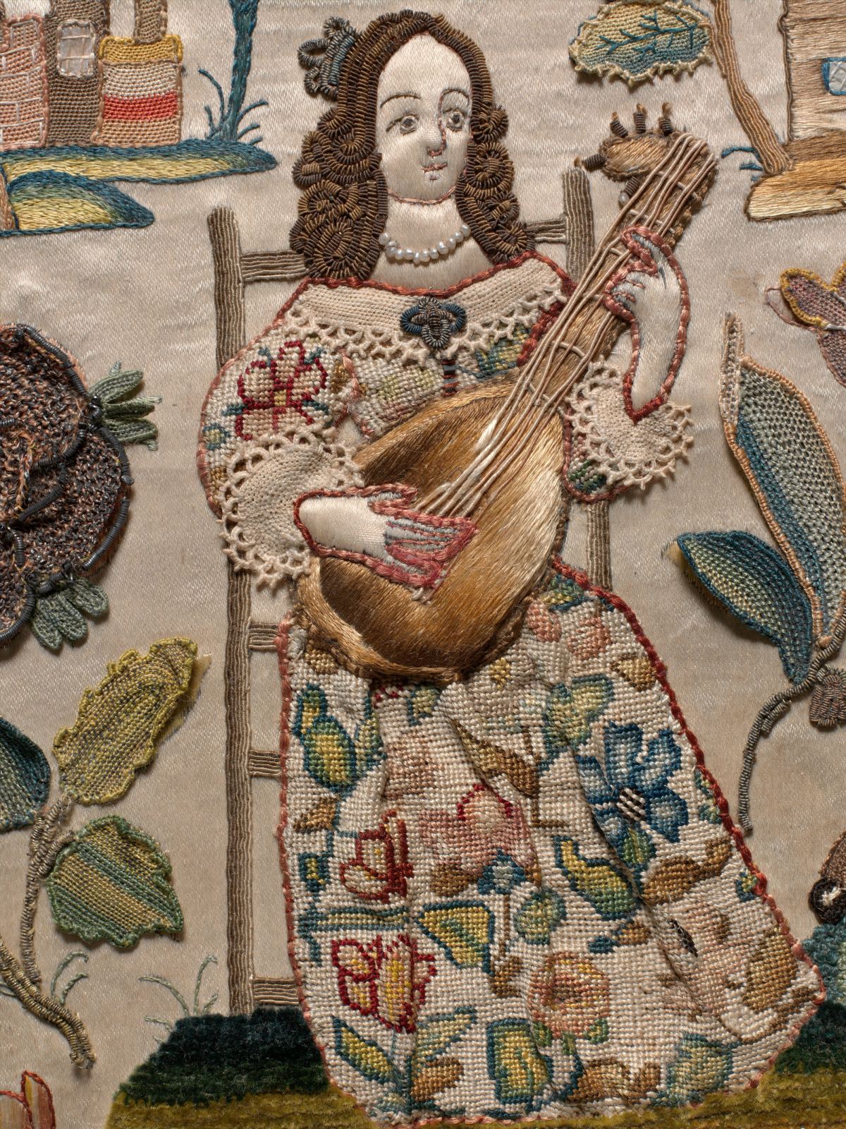 Close-up of an embroidered woman with coiled brown hair and a pearl necklace, seated on a ladder-back chair and playing a lute, in a lace-trimmed bodice and a skirt worked with flowers; raised leaves and flowers curl away from the cream satin at either side.