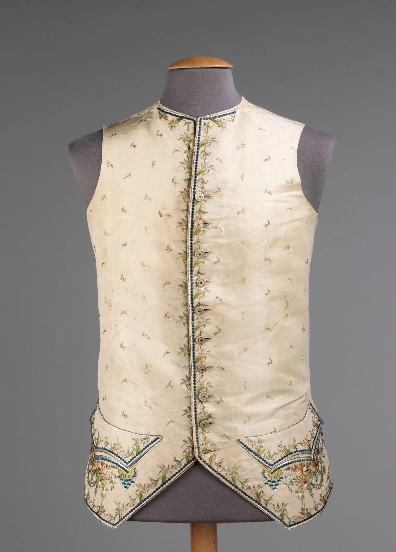 A cream silk waistcoat on a mannequin with a vertical row of small buttons down the center front, a narrow border of colored floral embroidery along the edge, and embroidered pocket flaps at the hem.