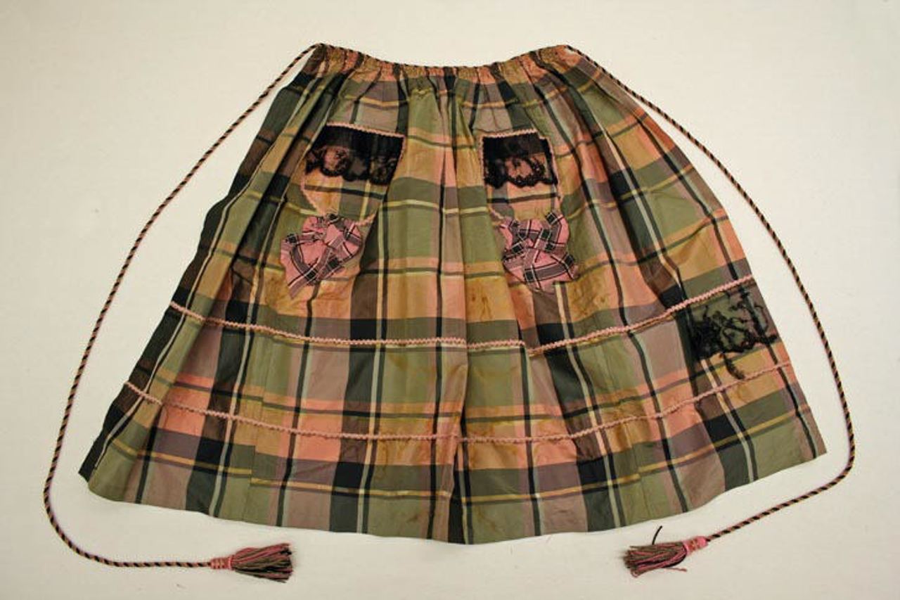 A pink, green and ochre plaid silk apron laid flat, gathered at the waist with long twisted cords ending in tassels, with two pockets trimmed with black lace and a plaid bow on each.