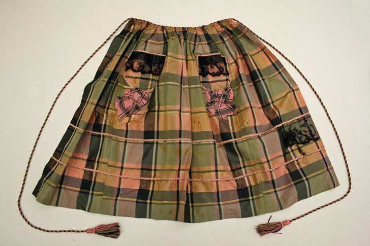 A pink, green and ochre plaid silk apron laid flat, gathered at the waist with long twisted cords ending in tassels, with two pockets trimmed with black lace and a plaid bow on each.