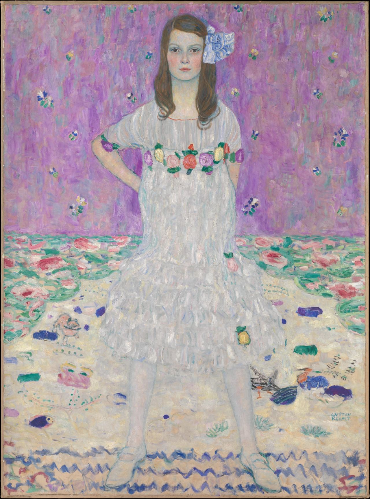 Full-length portrait of a girl with long brown hair and a pale blue bow, standing with her hands behind her hips on a patterned floor against a violet wall dotted with small flowers. She wears a white, loosely gathered dress with a band of bright roses across the chest and tiered ruffles at the skirt.