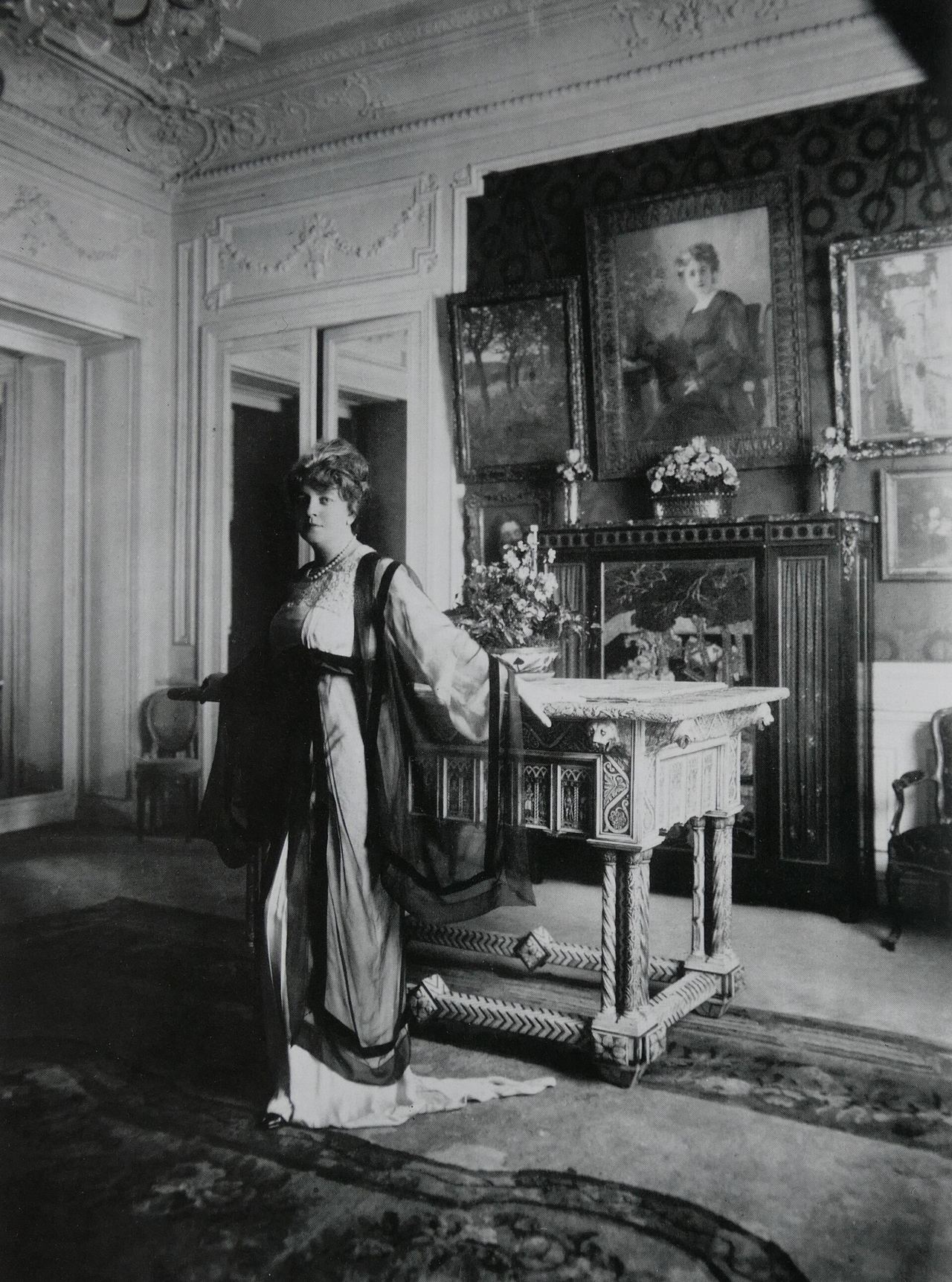 Black-and-white photograph of Jeanne Paquin standing in a paneled Paris drawing room, one hand resting on a carved stone-topped table with a vase of flowers. She wears a pale long dress under a sheer dark-edged overgown with wide sleeves and a pearl necklace; framed paintings hang behind her.