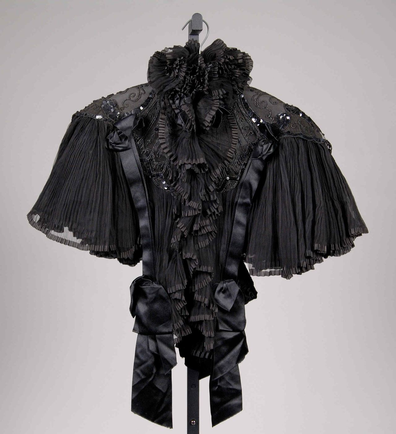 A short black evening cape on a stand, with pleated sheer sleeves, satin ribbon bows and a ruffled collar, and black sequins sewn over net on the shoulders.