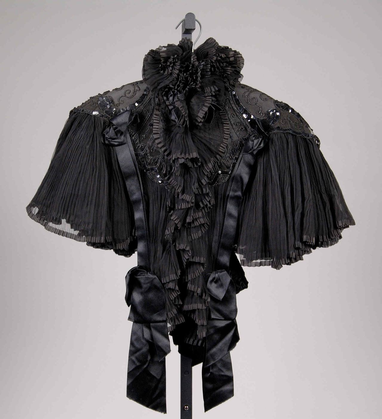 A short black evening cape on a stand, with pleated sheer sleeves, satin ribbon bows and a ruffled collar, and black sequins sewn over net on the shoulders.