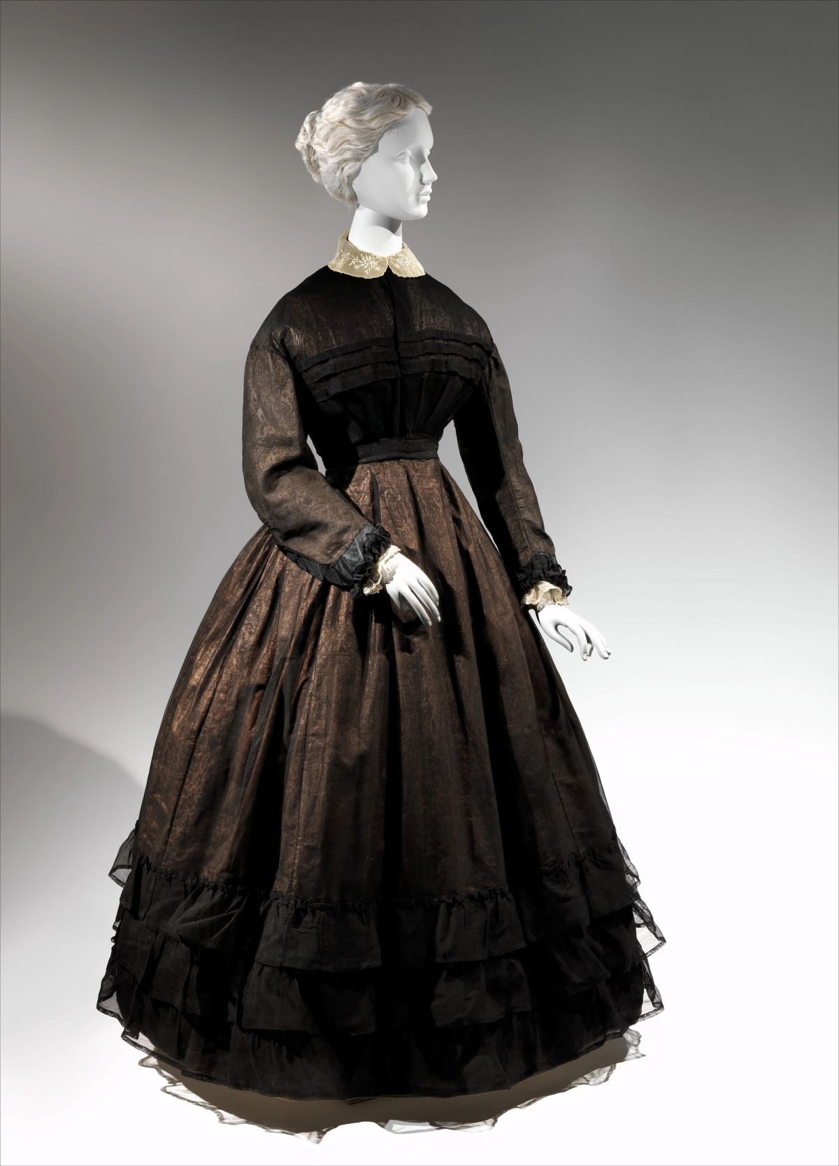 A woman's mourning dress of about 1867 on a mannequin: a full bell-shaped skirt of sheer black fabric over a brown-toned underlayer, tiered ruffles at the hem, pleated bodice, and a small cream collar and cuffs.
