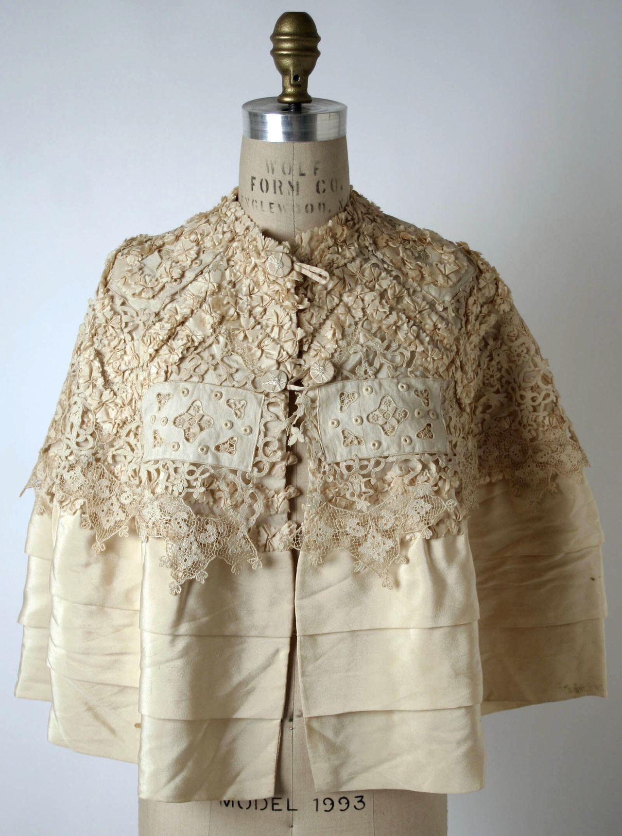 A short cream silk cape on a dress form, its collar and yoke covered in ivory lace motifs and its lower half falling in three pleated tiers of plain silk, fastened at the front with small loops.