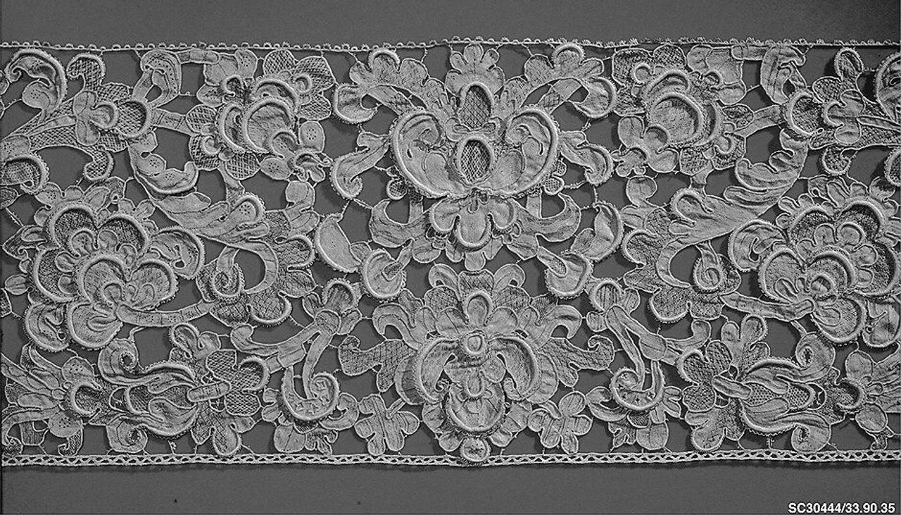 Black-and-white photograph of a wide border of heavy needle lace: padded, raised scrolls and flowers, with small net-like patches inside the shapes, joined by bars and with a small beaded edge along both sides.