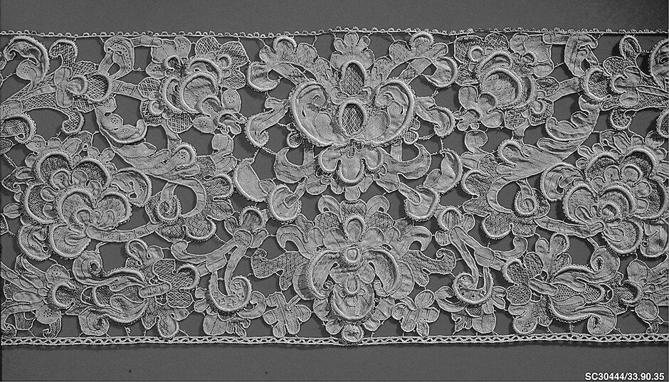 Black-and-white photograph of a wide border of heavy needle lace: padded, raised scrolls and flowers, with small net-like patches inside the shapes, joined by bars and with a small beaded edge along both sides.