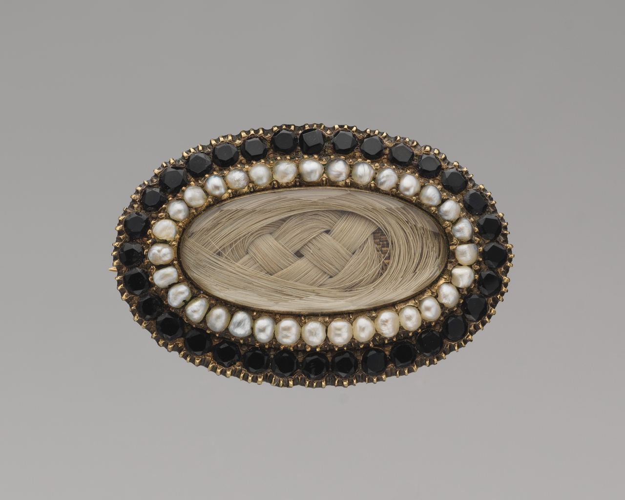 An oval gold brooch with a braided panel of light hair under glass, ringed by seed pearls and an outer ring of faceted black stones.