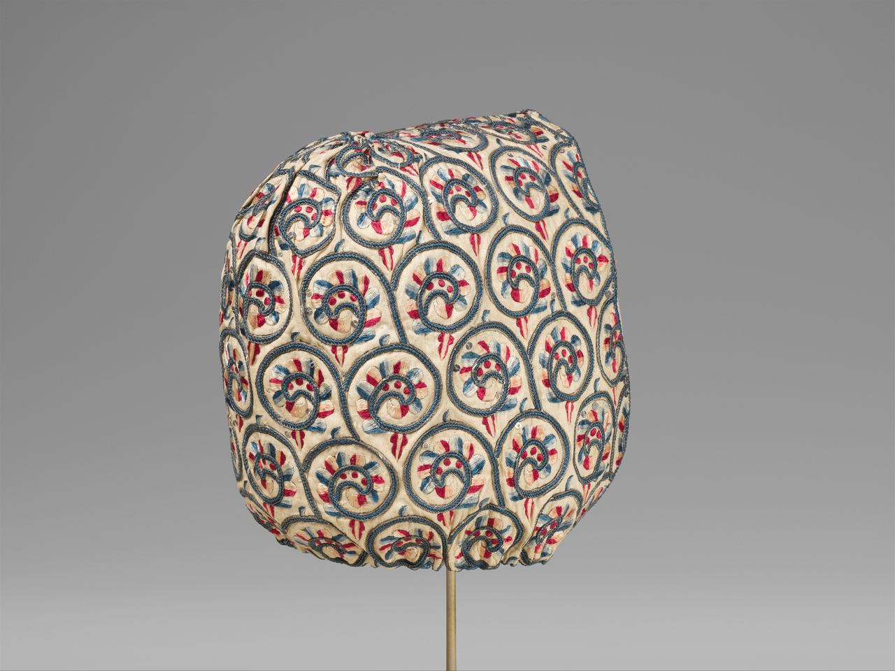 A linen coif on a stand, embroidered all over with scrolling vines and flowers in blue, red and pink silk, with tiny metal spangles catching the light between the scrolls.