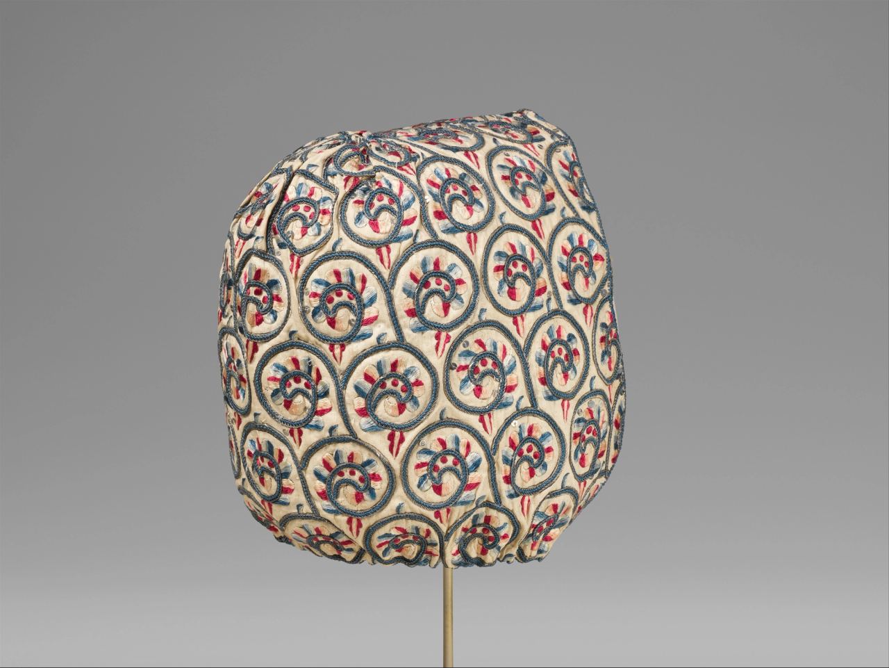 A linen coif on a stand, embroidered all over with scrolling vines and flowers in blue, red and pink silk, with tiny metal spangles catching the light between the scrolls.