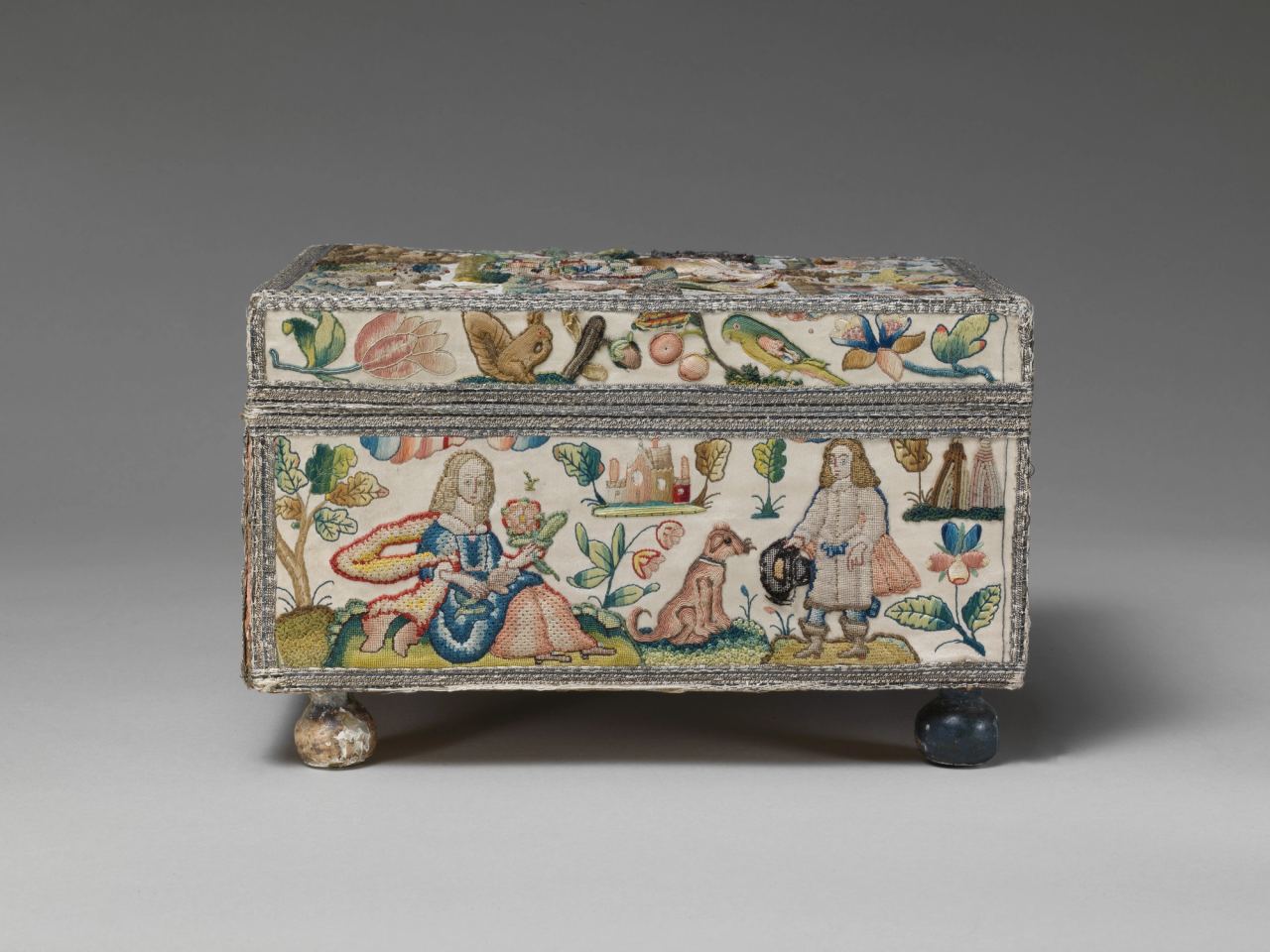 A small rectangular casket covered in cream satin, seen from the side. Embroidered panels show a seated woman holding a flower, a dog, a man in a wide-brimmed hat with a cloak, a castle, a squirrel, a parrot and flowers, framed by silver metal-thread braid, on two round turned feet.