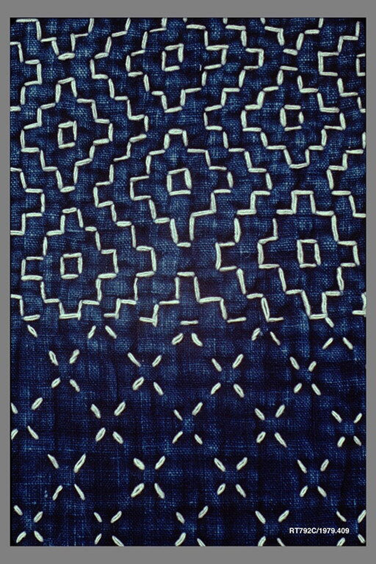 Close view of indigo cloth stitched with white thread: above, rows of running stitches turn at right angles to outline stepped diamonds with open squares at their centers; below, small crosses each made from four short, separate stitches.