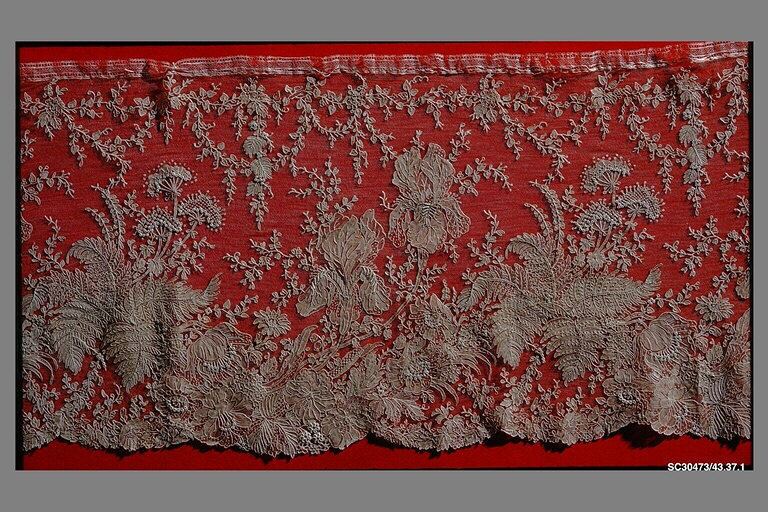 A length of cream needle lace pinned over red cloth, with a scalloped lower edge and a design of large irises, ferns, grape-like leaves and sprays of small flowers over a fine net ground.