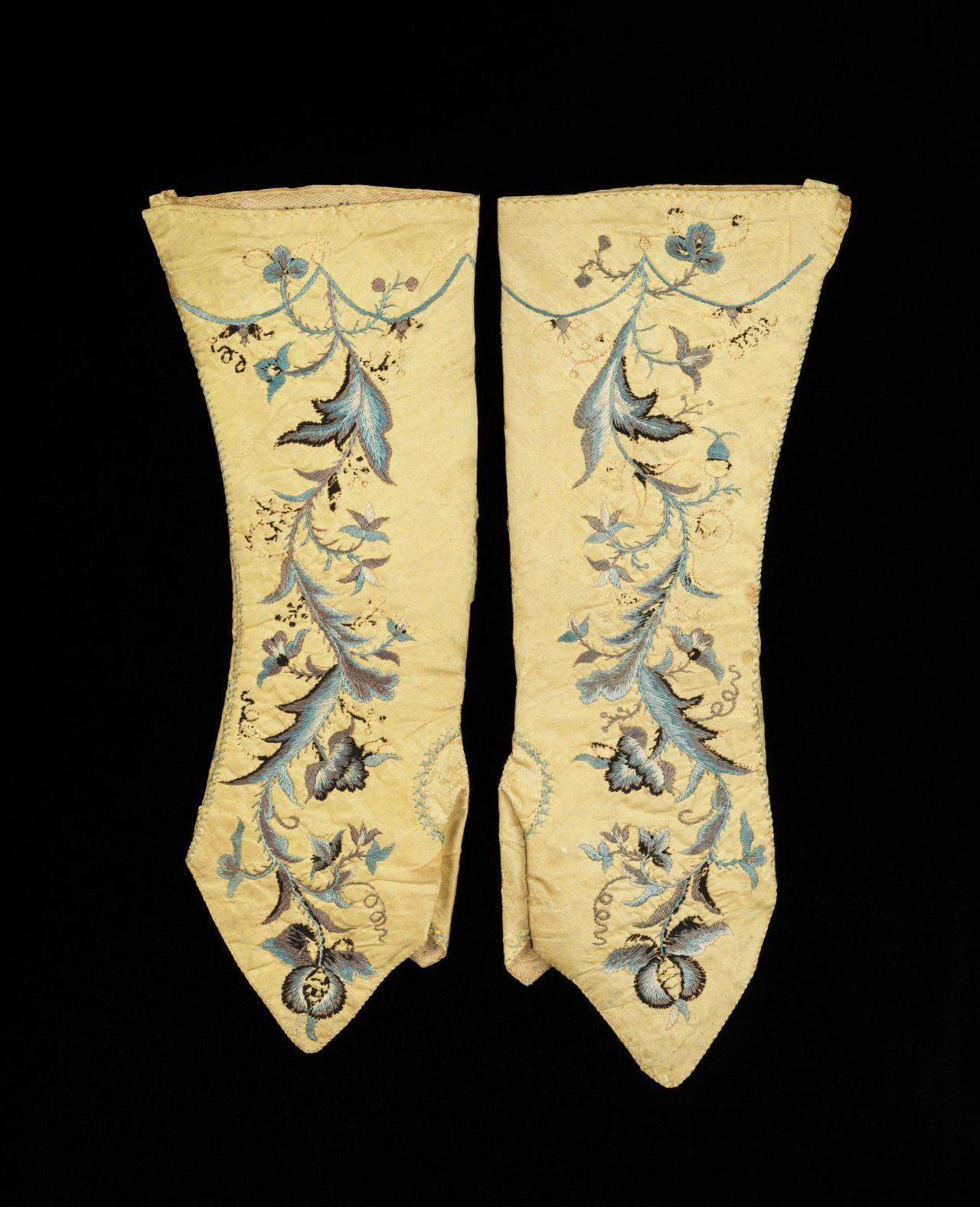 A pair of pale yellow silk mitts laid flat on black, each with a pointed flap at the hand end and a trailing spray of blue and grey leaves, flowers and tendrils embroidered up its length.