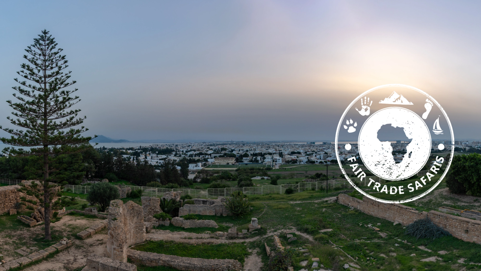 View over historic stone ruins and green grounds overlooking a coastal city at sunset, with the Fair Trade Safaris logo displayed on the right.