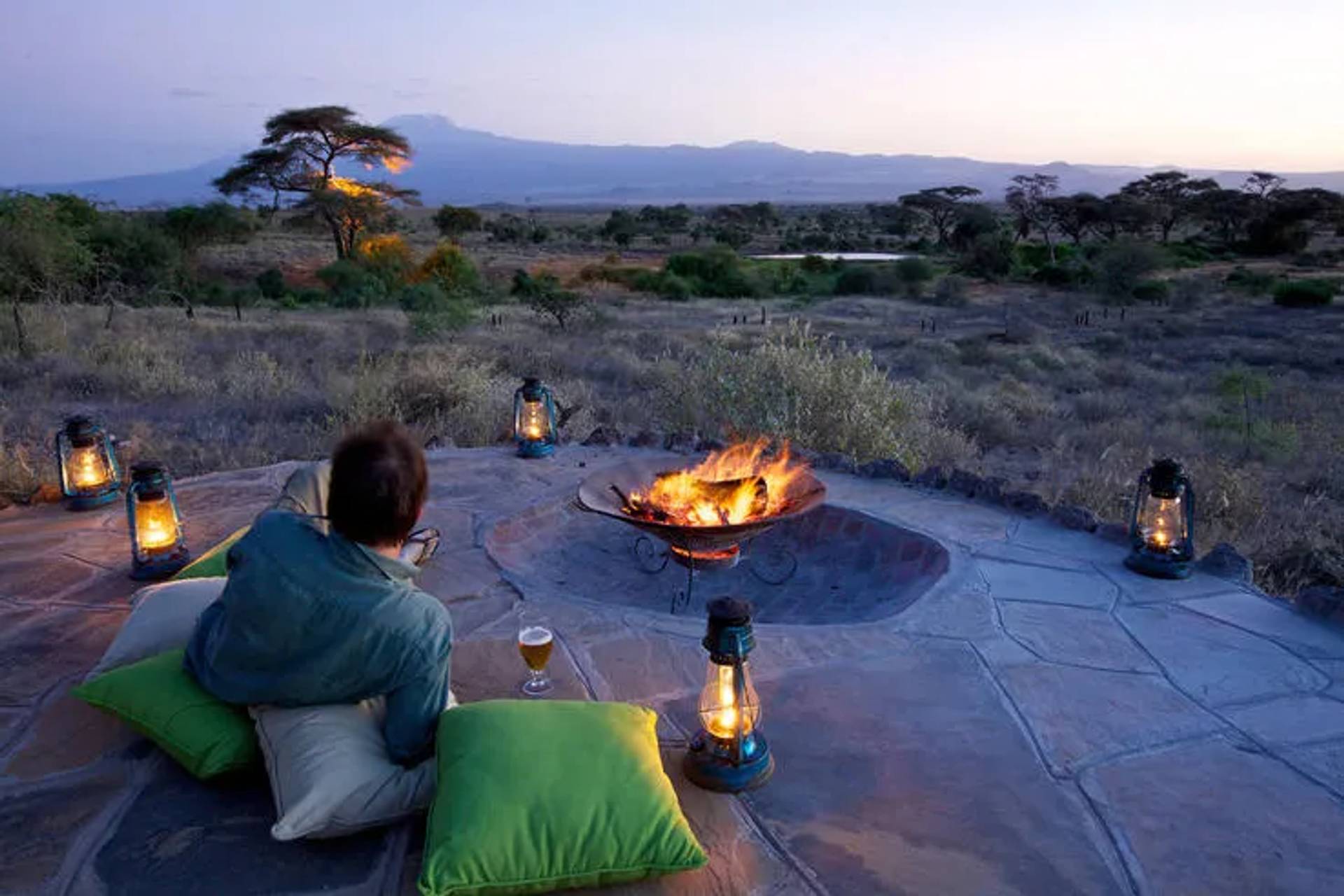 Person relaxing by a fire pit with lanterns at sunset, overlooking African wilderness and distant mountains