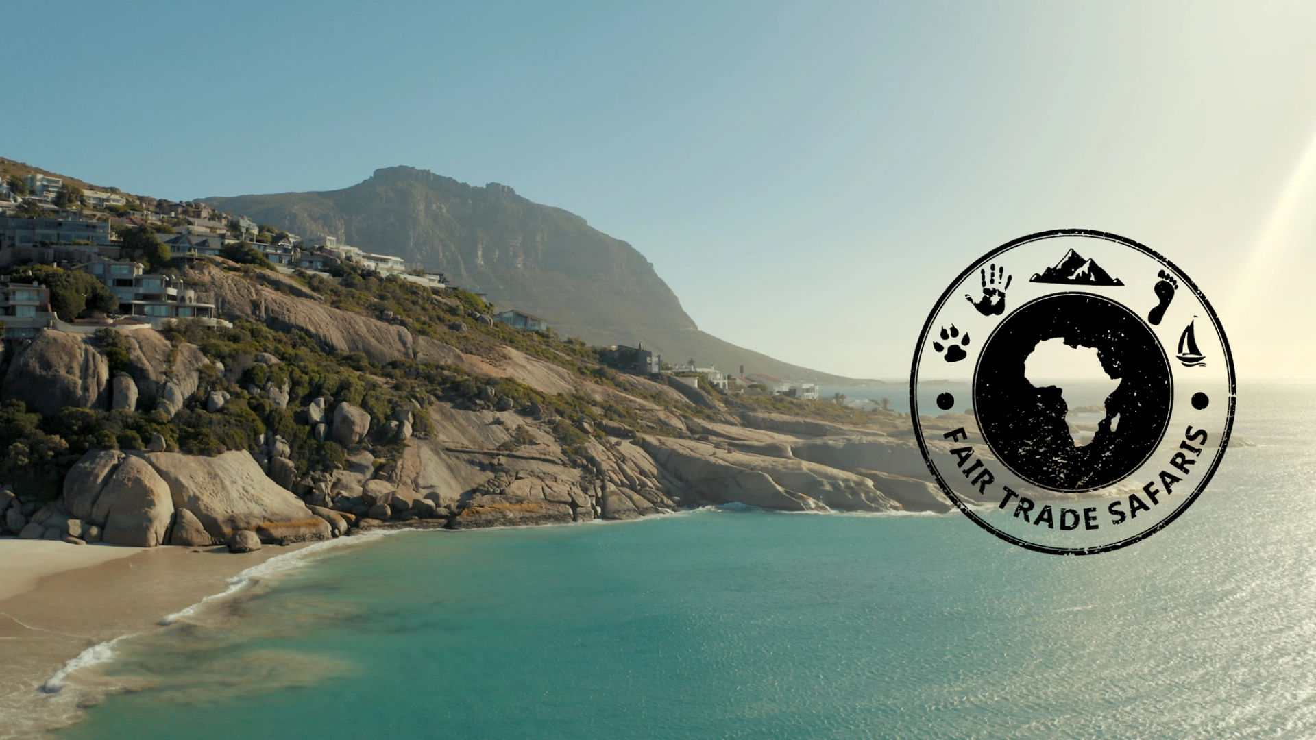 Turquoise ocean along a rocky coastline with hillside homes and a mountain backdrop, featuring the Fair Trade Safaris logo on the right.