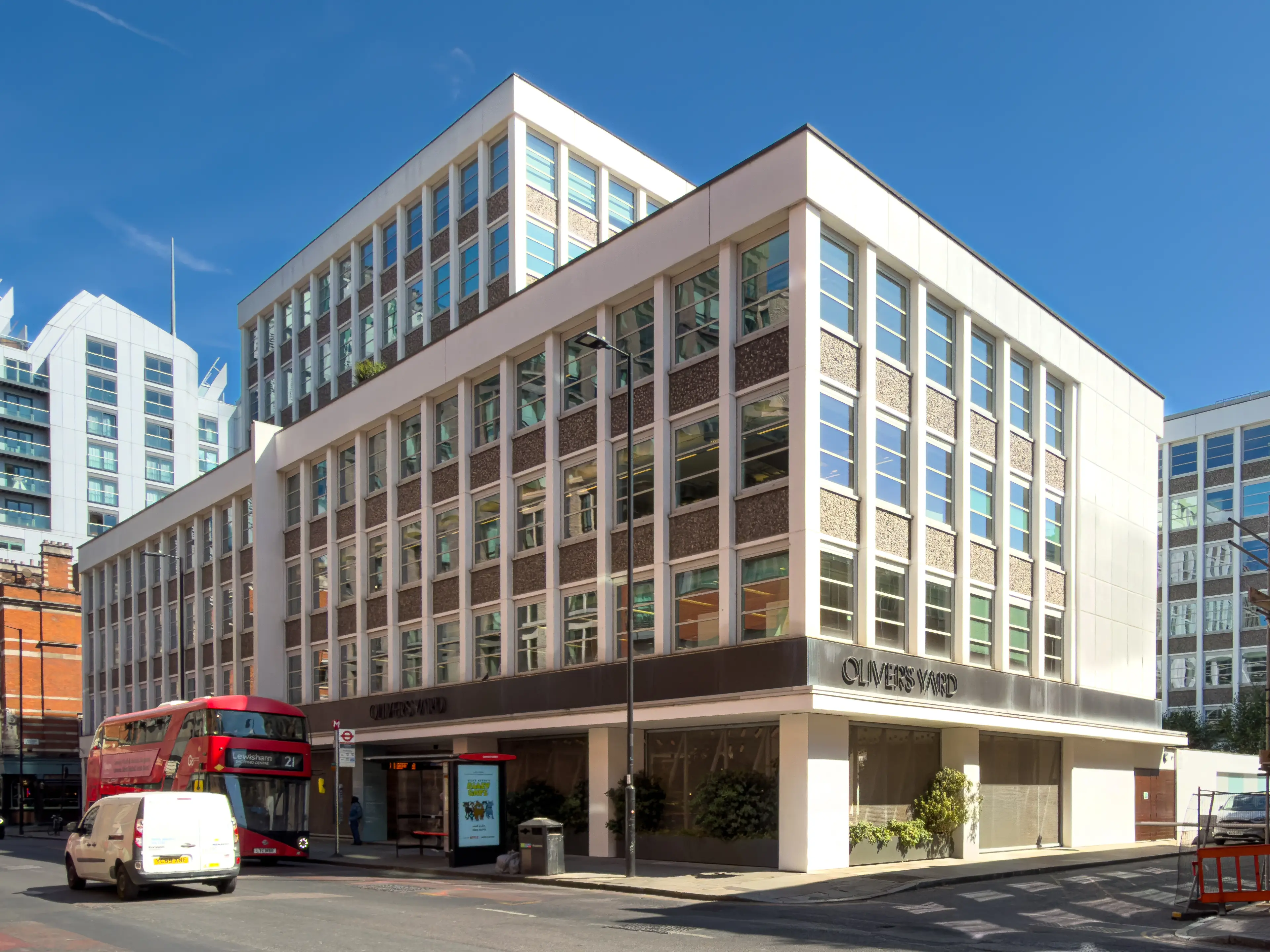 Exterior of the multi-story Oliver's Yard building on a sunny street, with a red double-decker bus and white van.