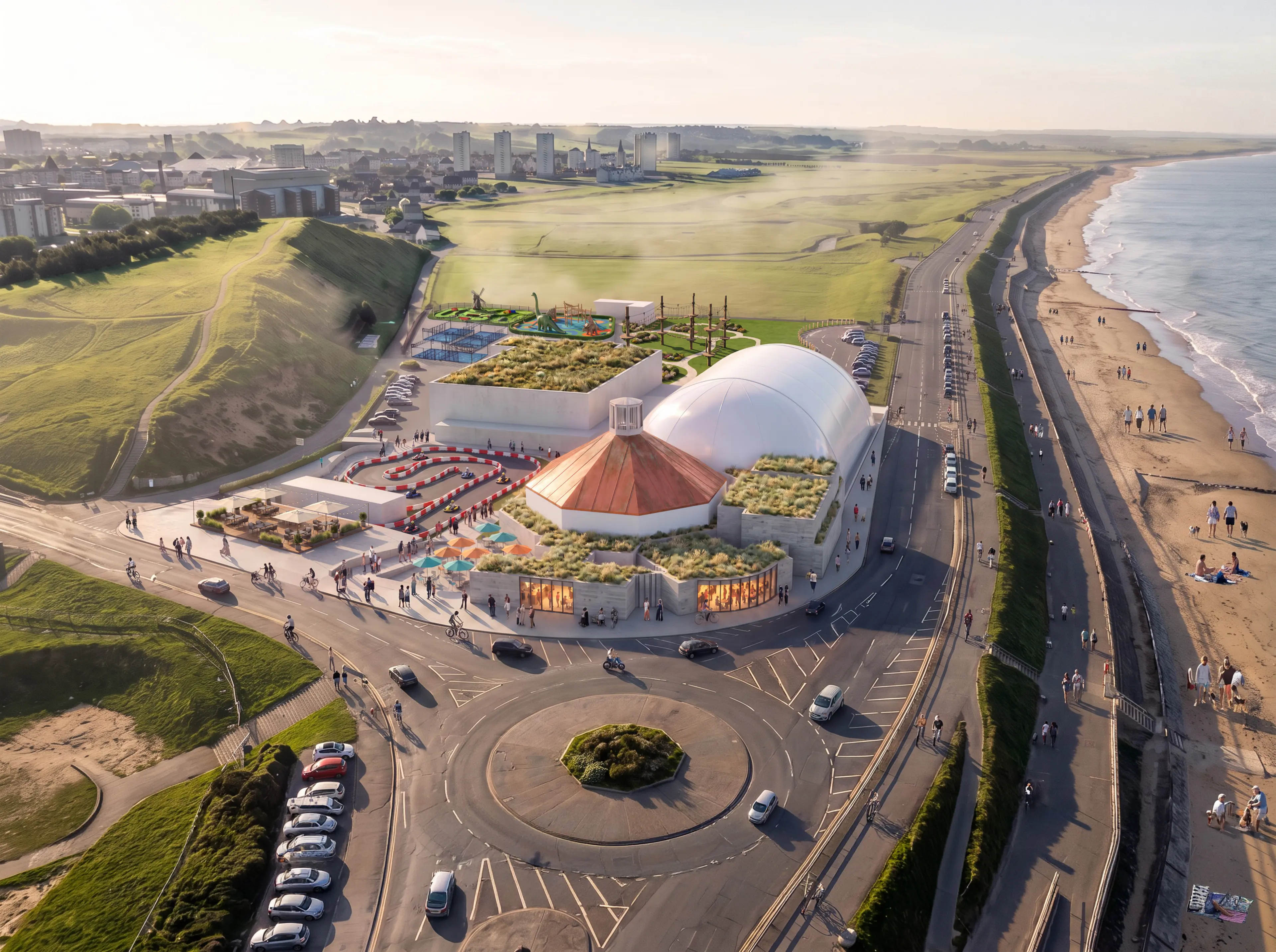 Aerial view of a modern coastal complex with green roofs and a copper dome, situated beside a sandy beach and a road.