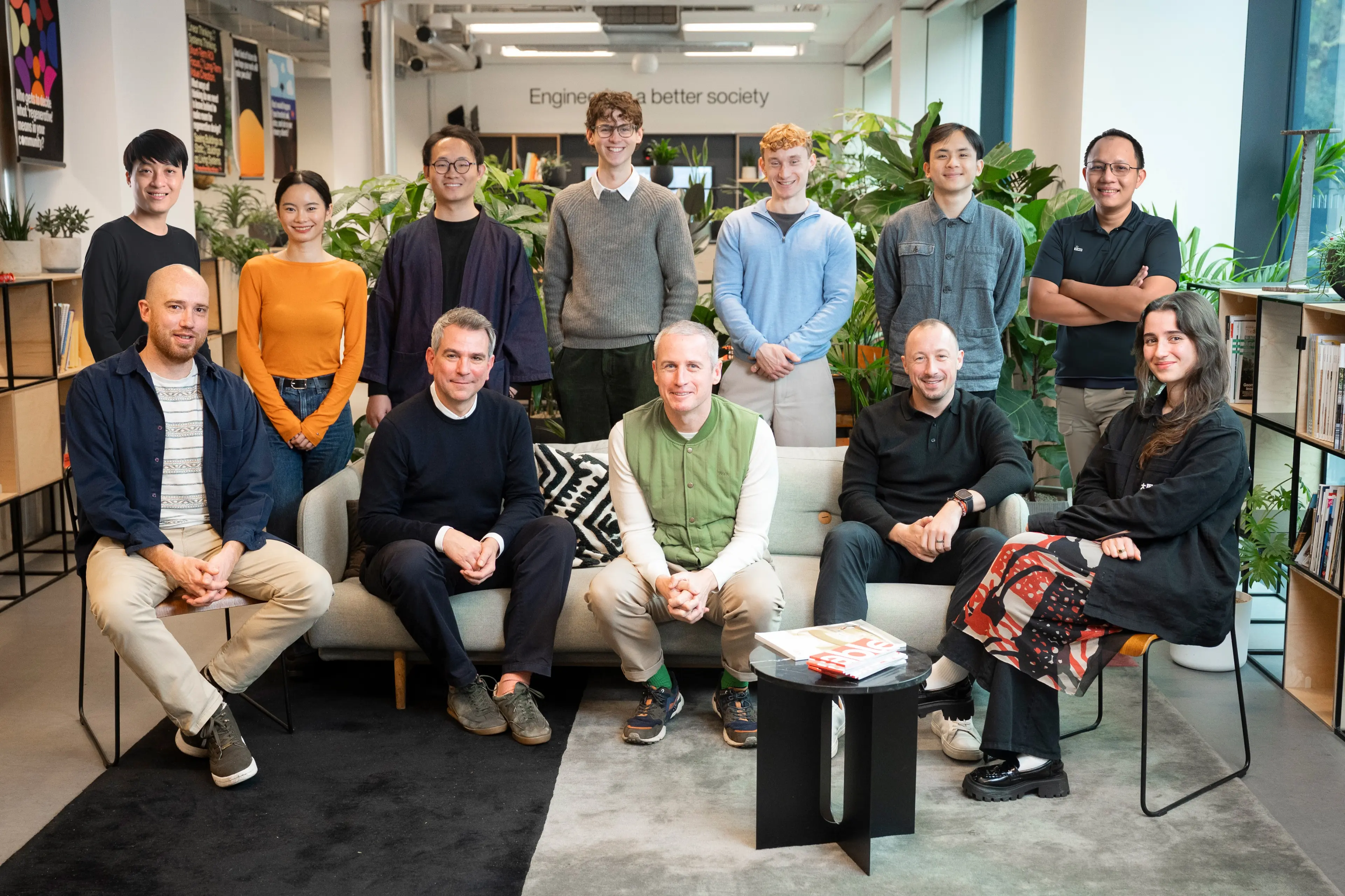 A diverse group of 13 people, some standing and some sitting, posing in a modern, plant-filled office.