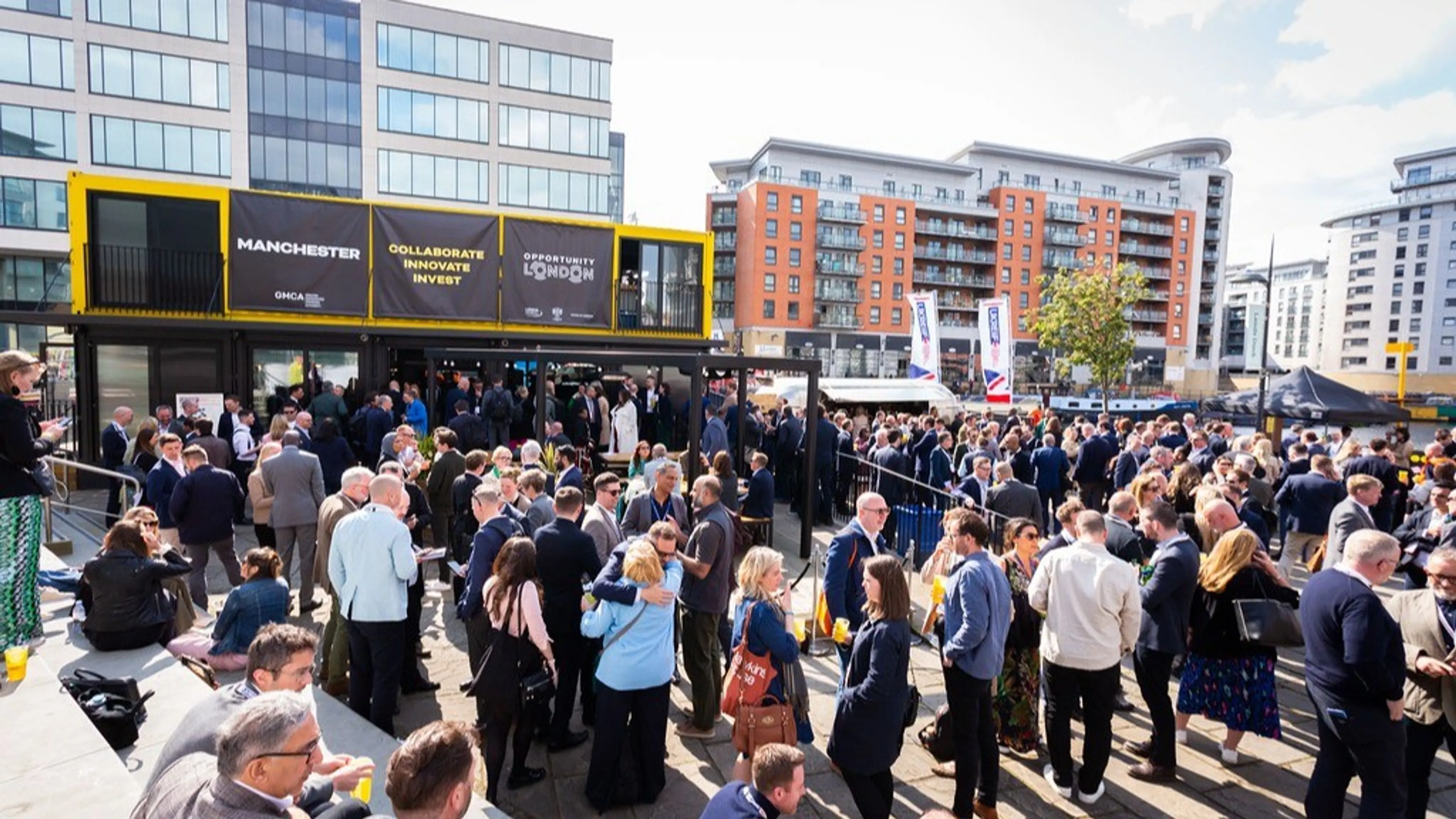 An outdoor networking event with a large crowd in front of yellow containers promoting investment in Manchester and London.