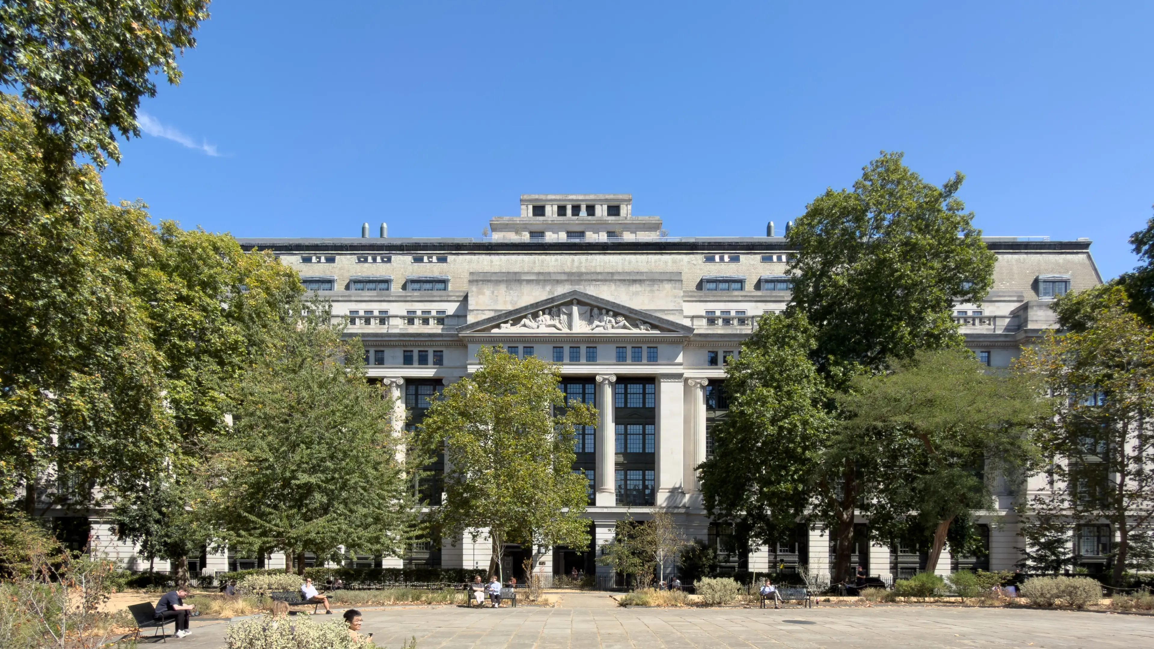 A grand classical building with columns and a pediment, partially obscured by green trees, with people relaxing in a park below.