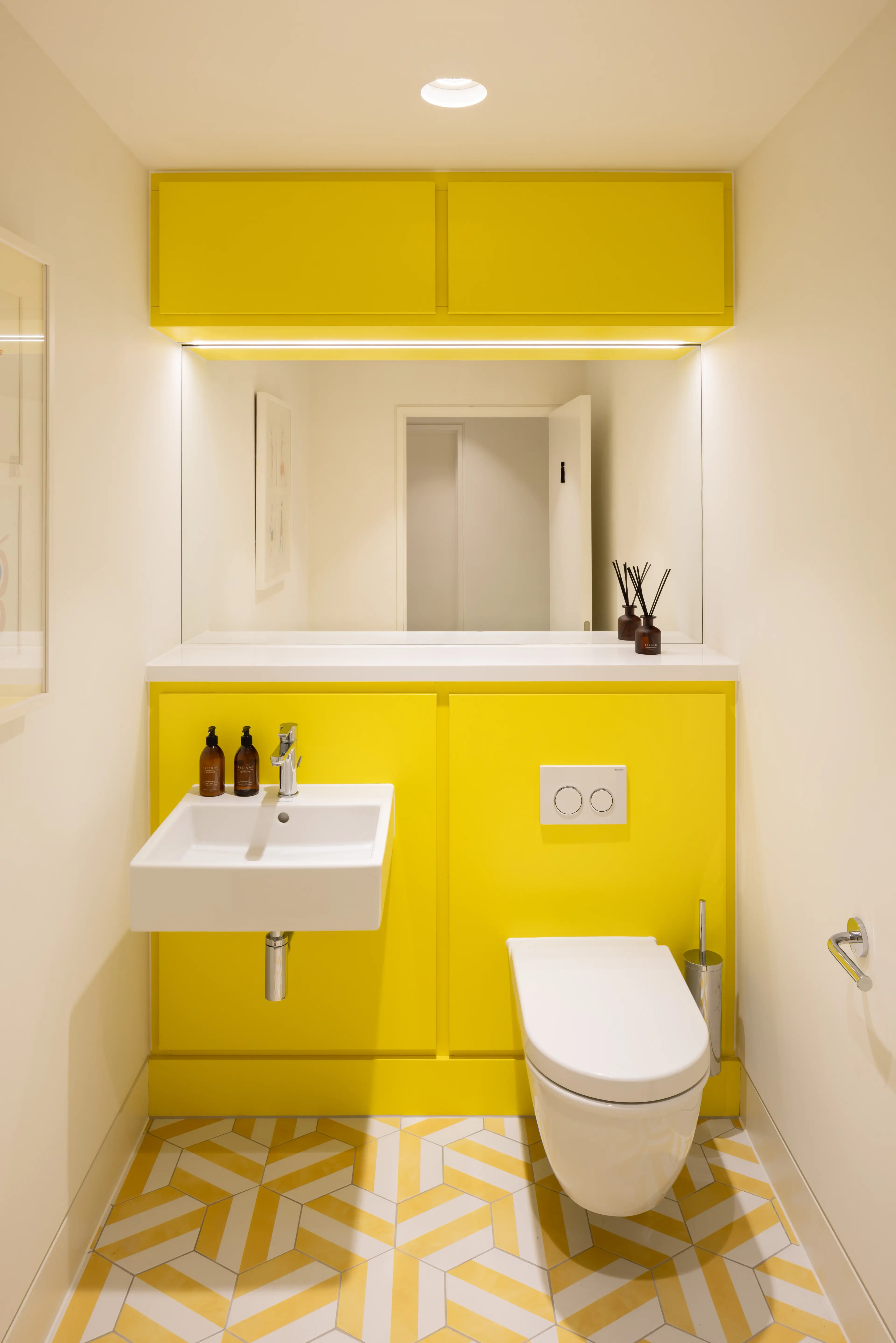 A modern bathroom with bright yellow lower walls and upper cabinets, a white sink, a wall-mounted toilet, and a yellow and white geometric tiled floor.