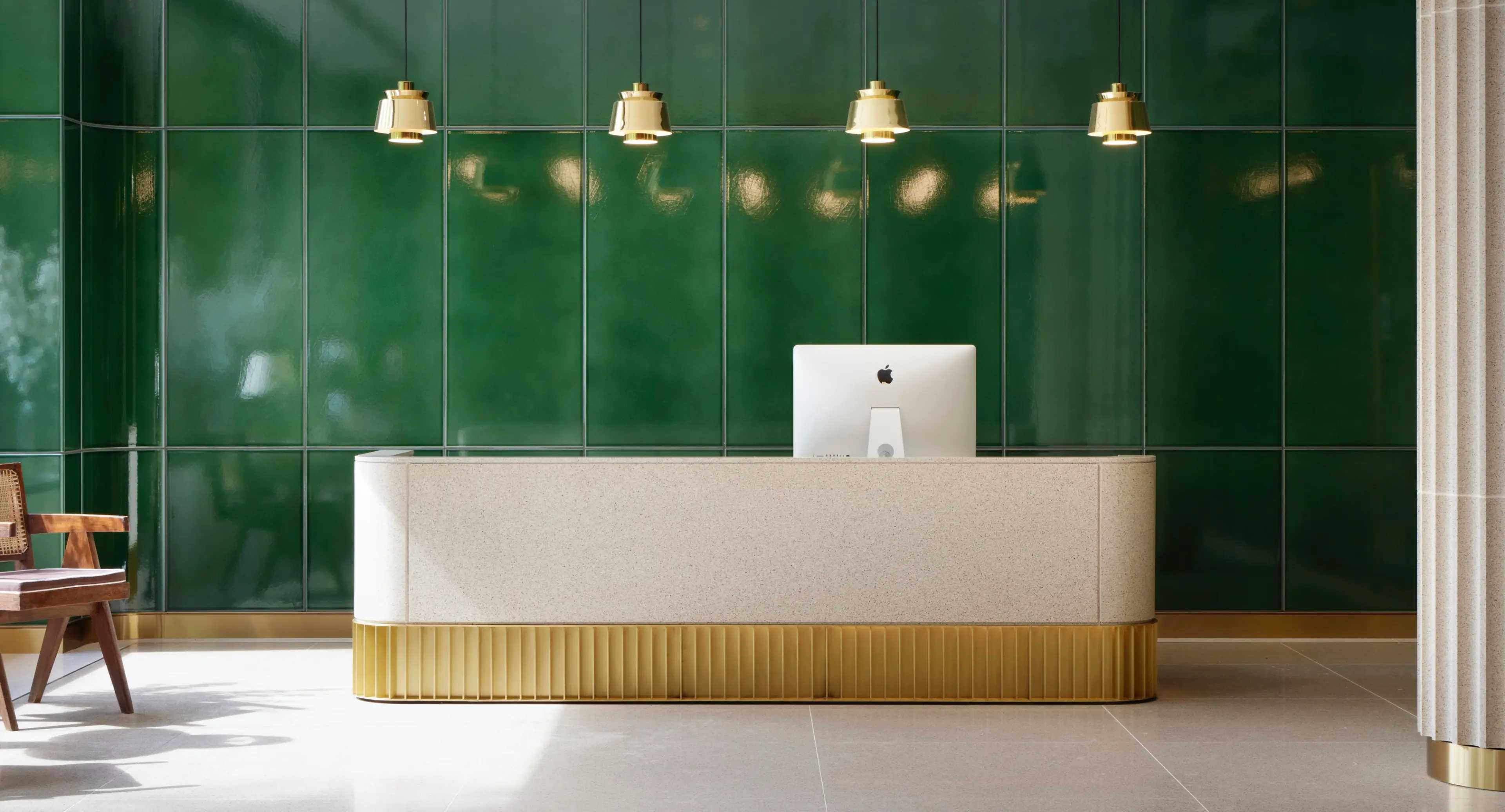 A modern, curved reception desk with an iMac and gold fluted base, set against a dark green tiled wall under gold pendant lights.