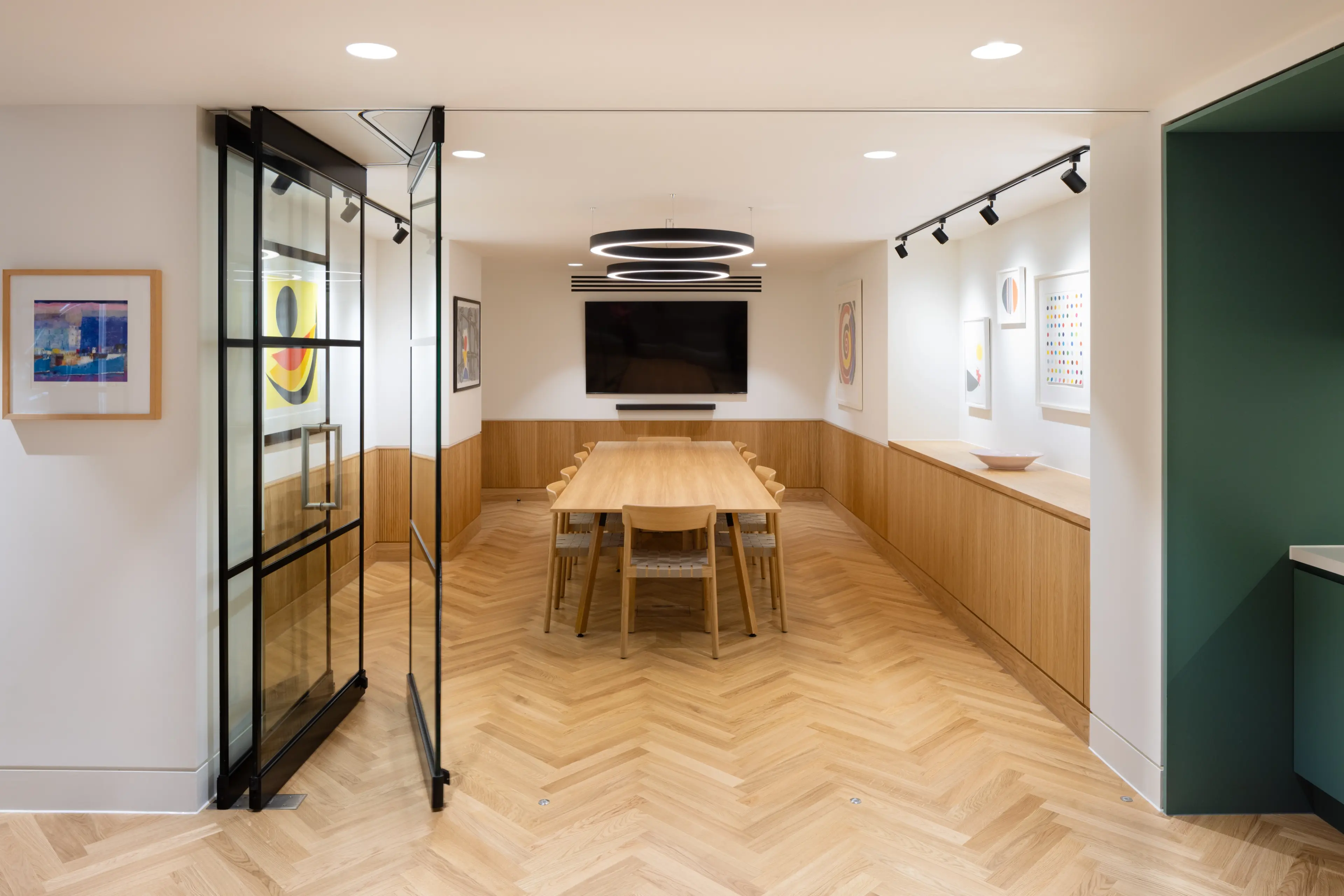 A modern conference room with a long wooden table, chairs, a large TV screen, herringbone wood floor, and an open glass door.