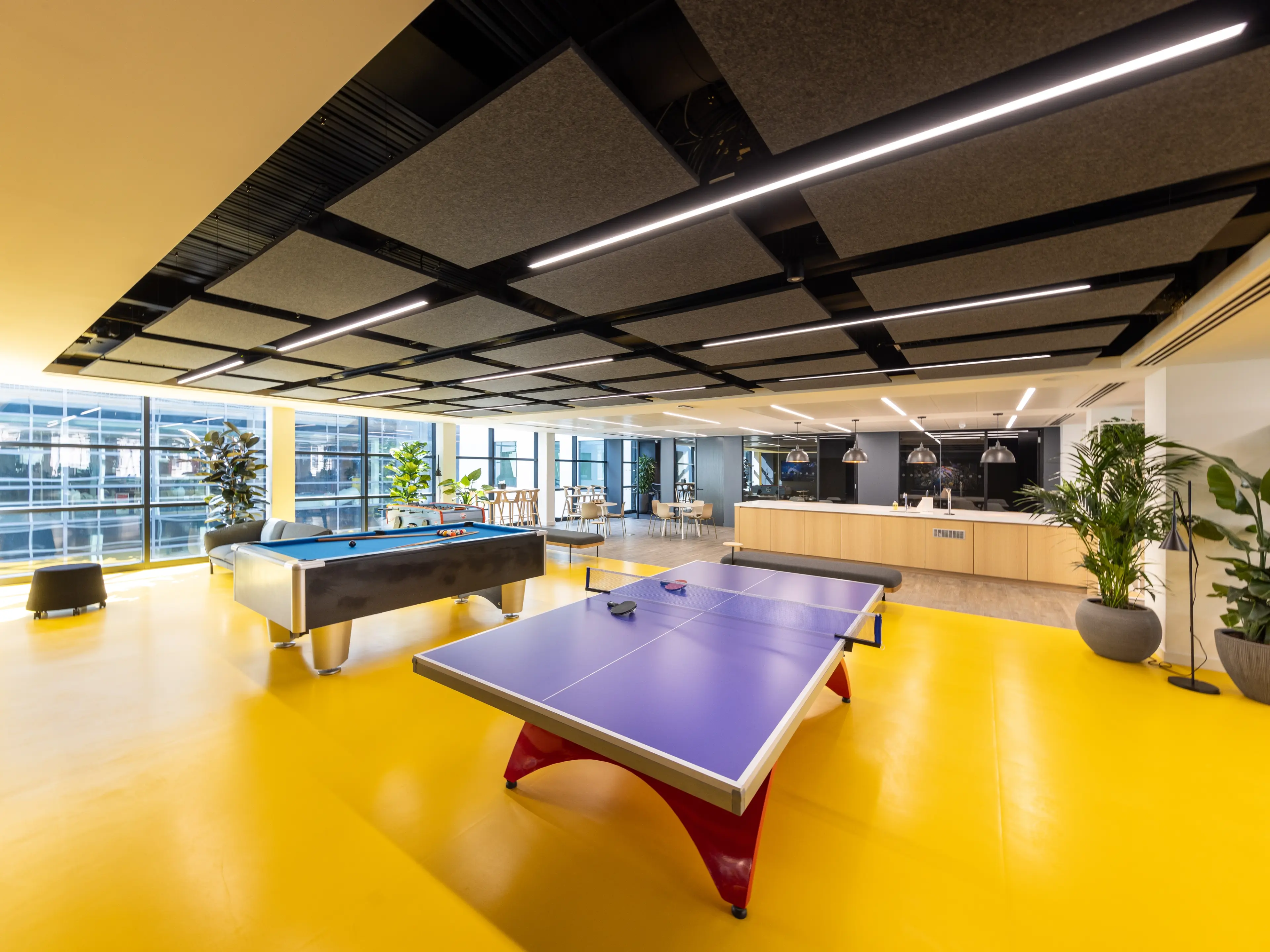 A vibrant, modern office break room featuring a yellow floor, a pool table, a ping-pong table, large windows, and a kitchenette.