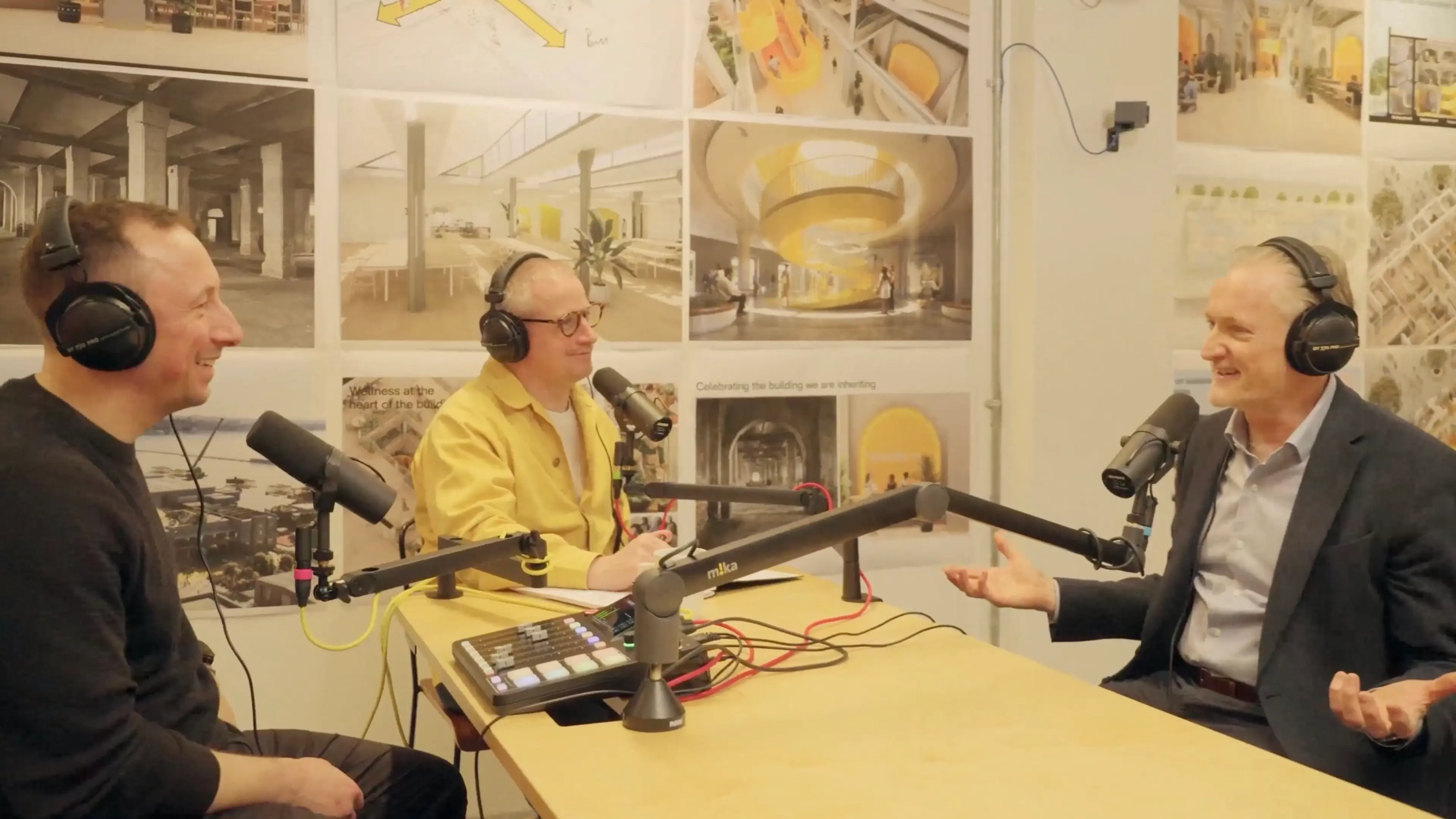 Three men recording a podcast, smiling and talking, with architectural plans covering the wall behind them.