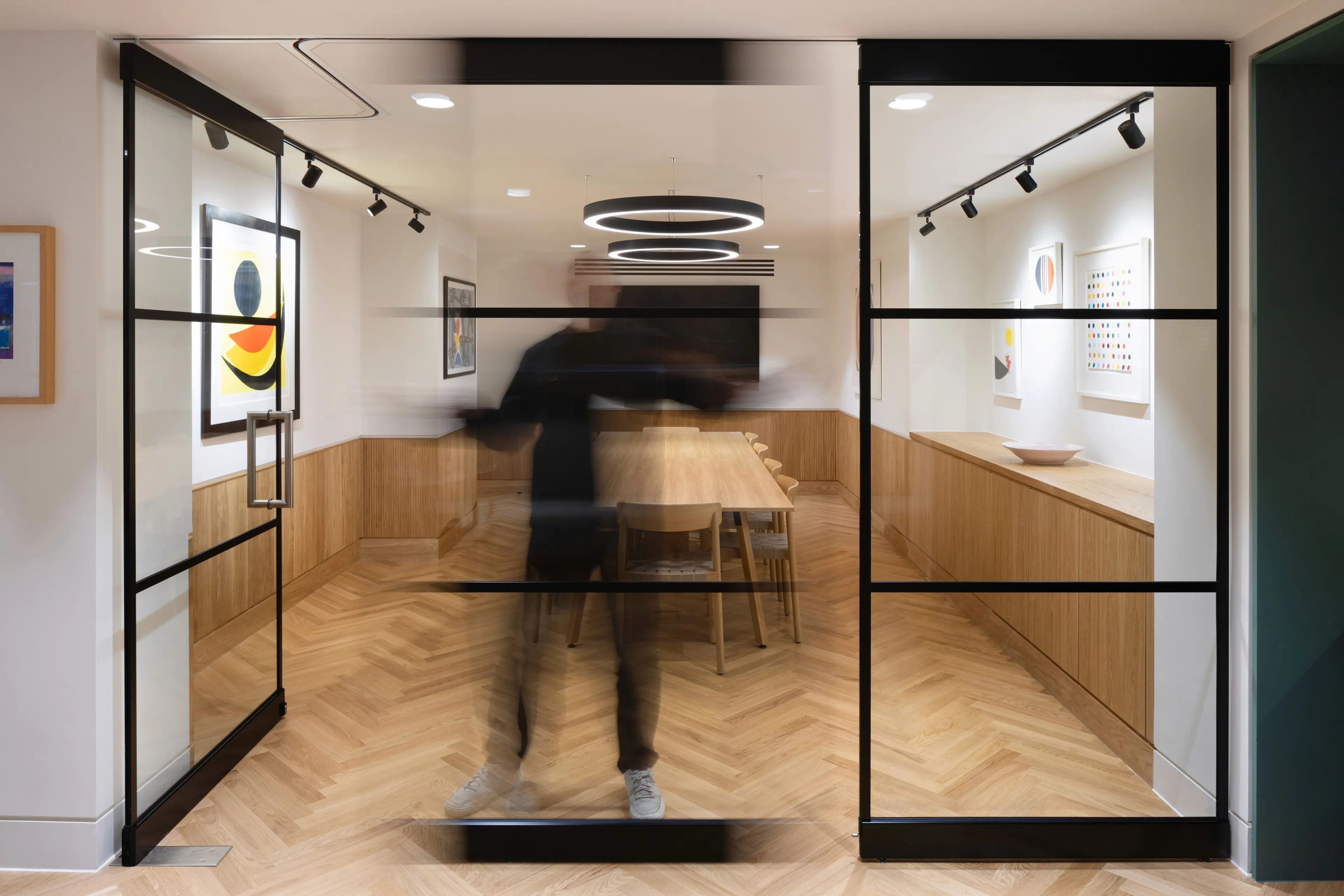 A modern meeting room with a person in motion blur passing through glass doors, featuring a herringbone wood floor and wooden furnishings.