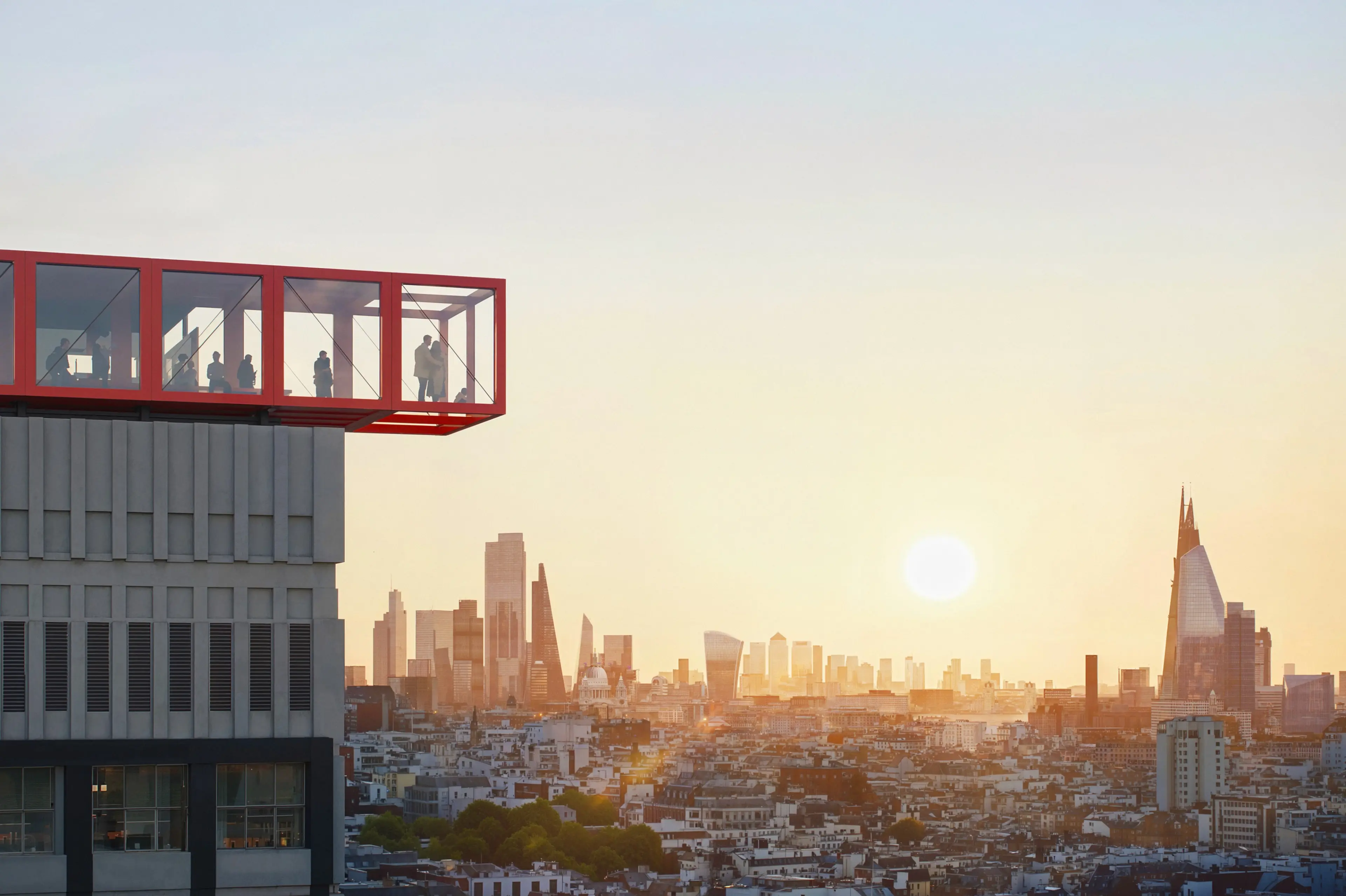 Red cantilevered glass observation deck with people overlooking a city skyline at sunset.