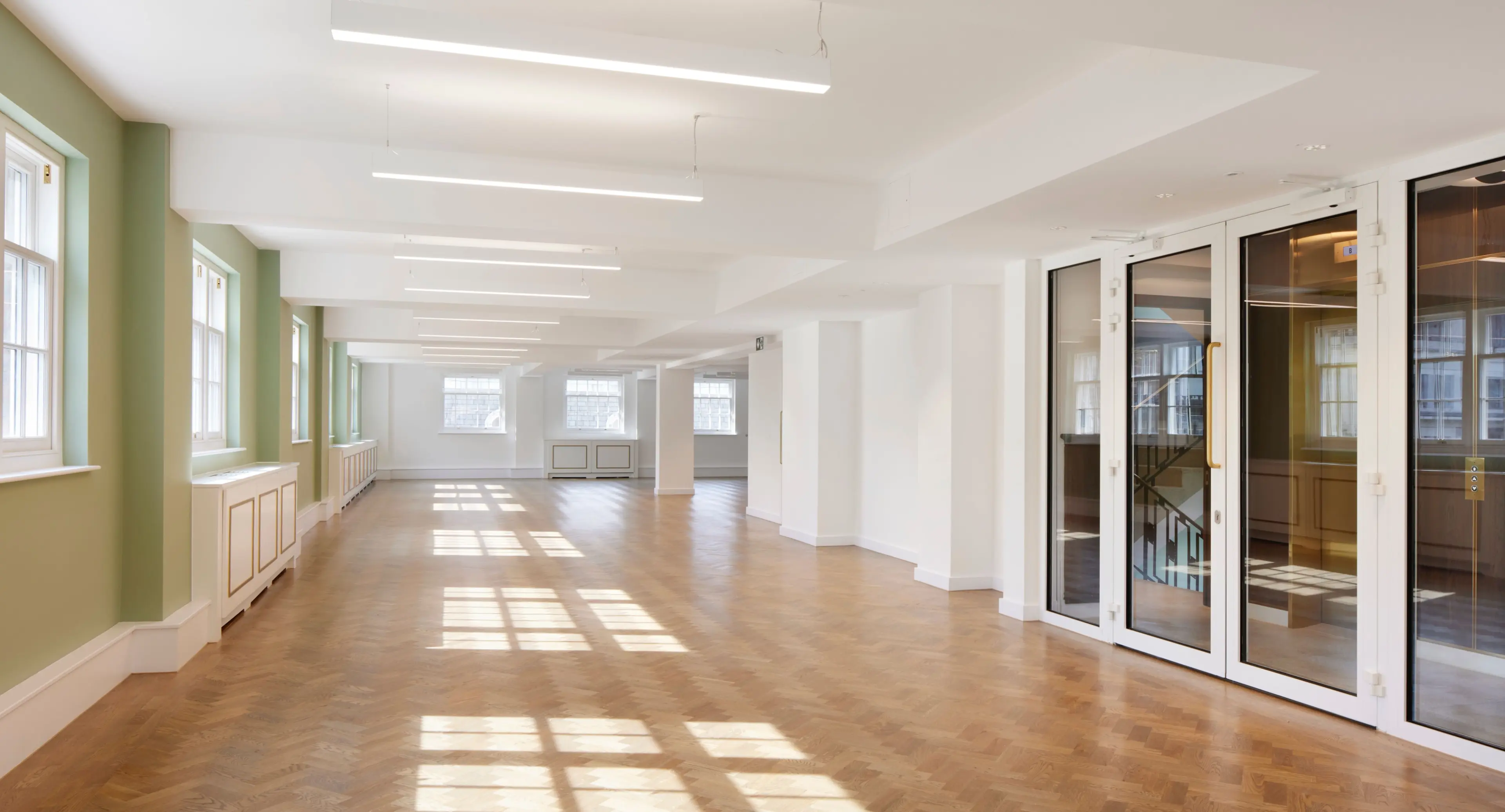 An empty, sunlit room with light green walls, a parquet floor, and large glass doors.