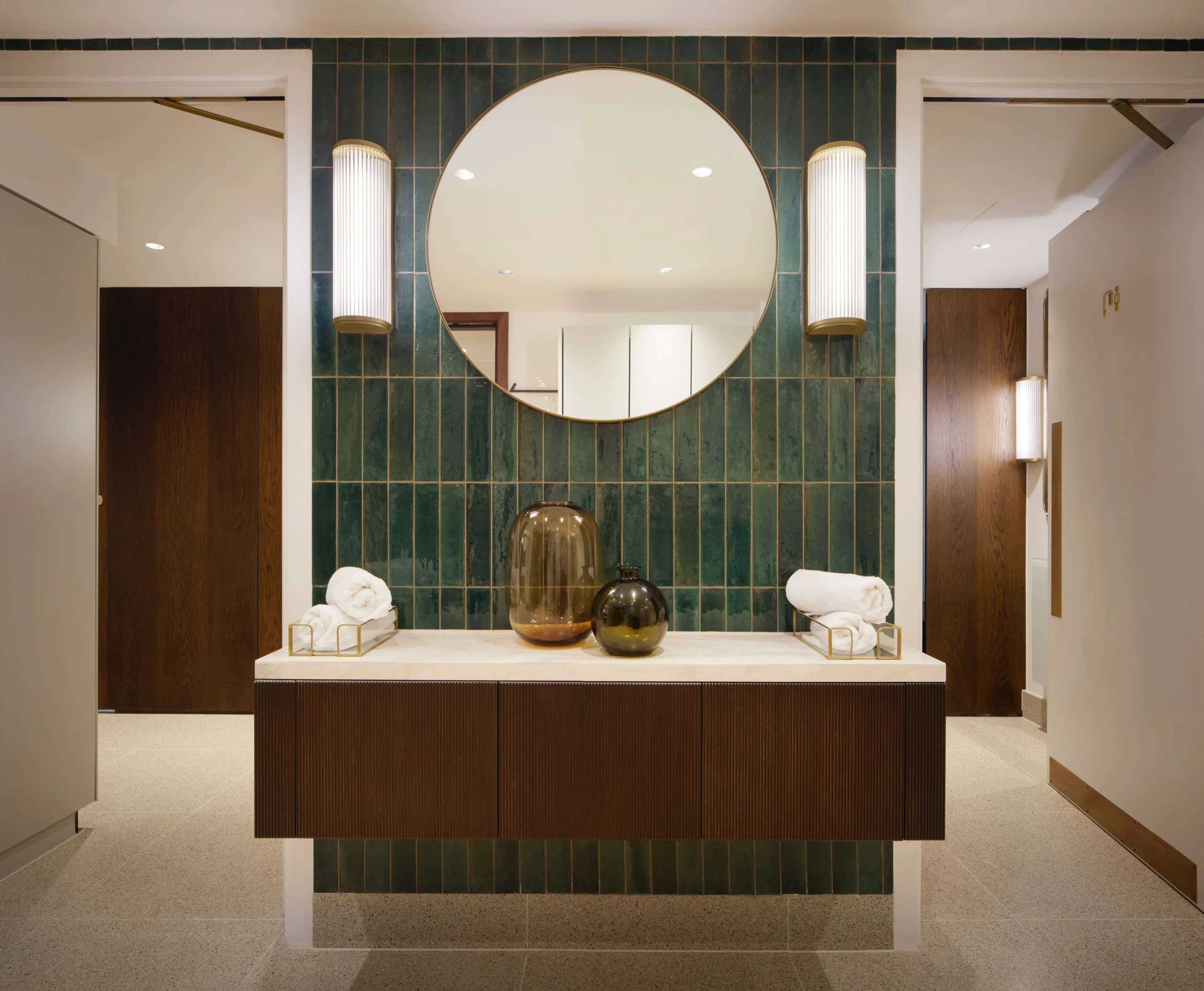 A modern vanity with a round mirror, dark green tiled wall, and dark wood cabinet, flanked by two doorways.